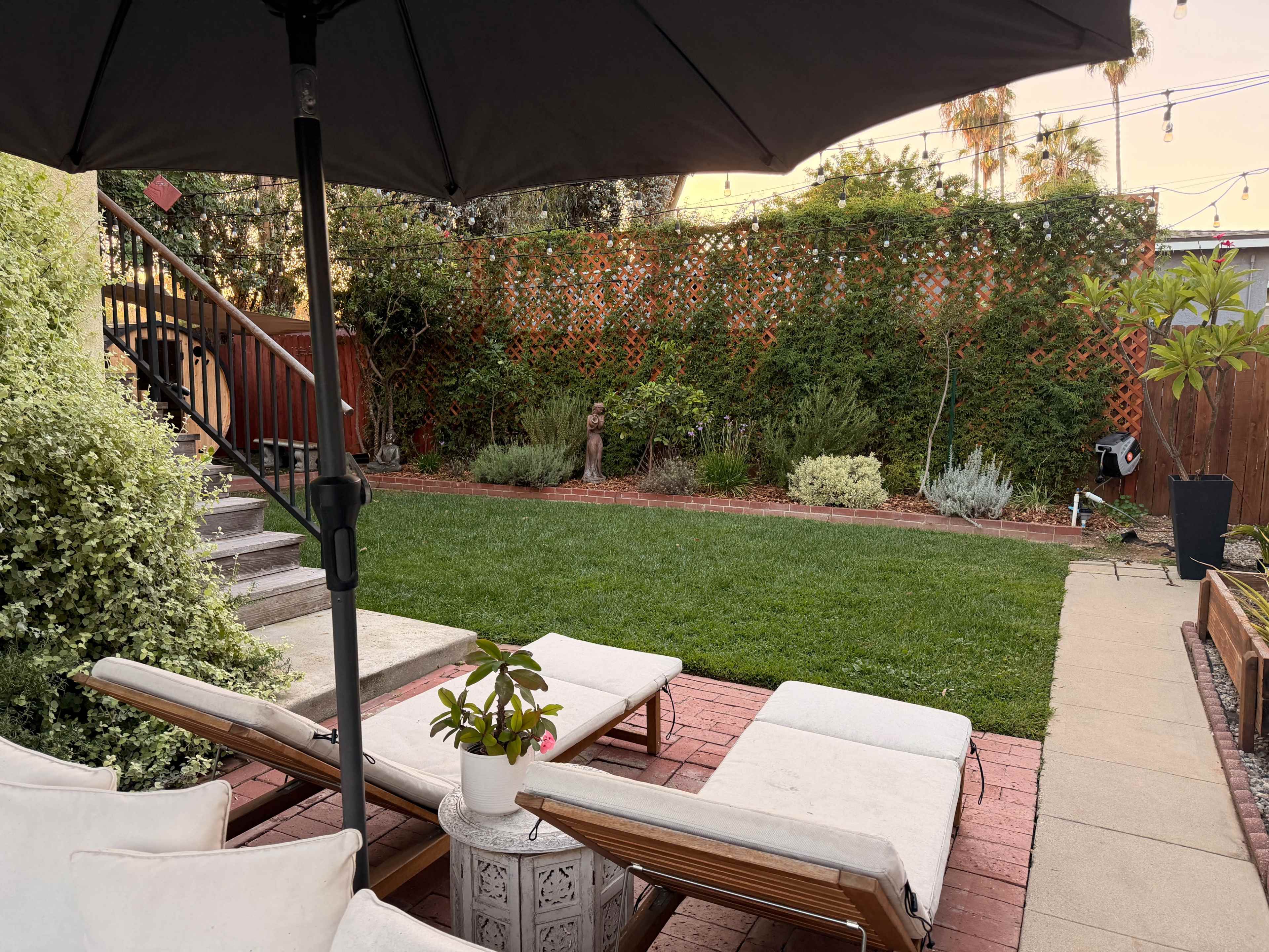 The image shows a well-maintained backyard with a lawn, surrounded by plants and outdoor furniture beneath an umbrella.