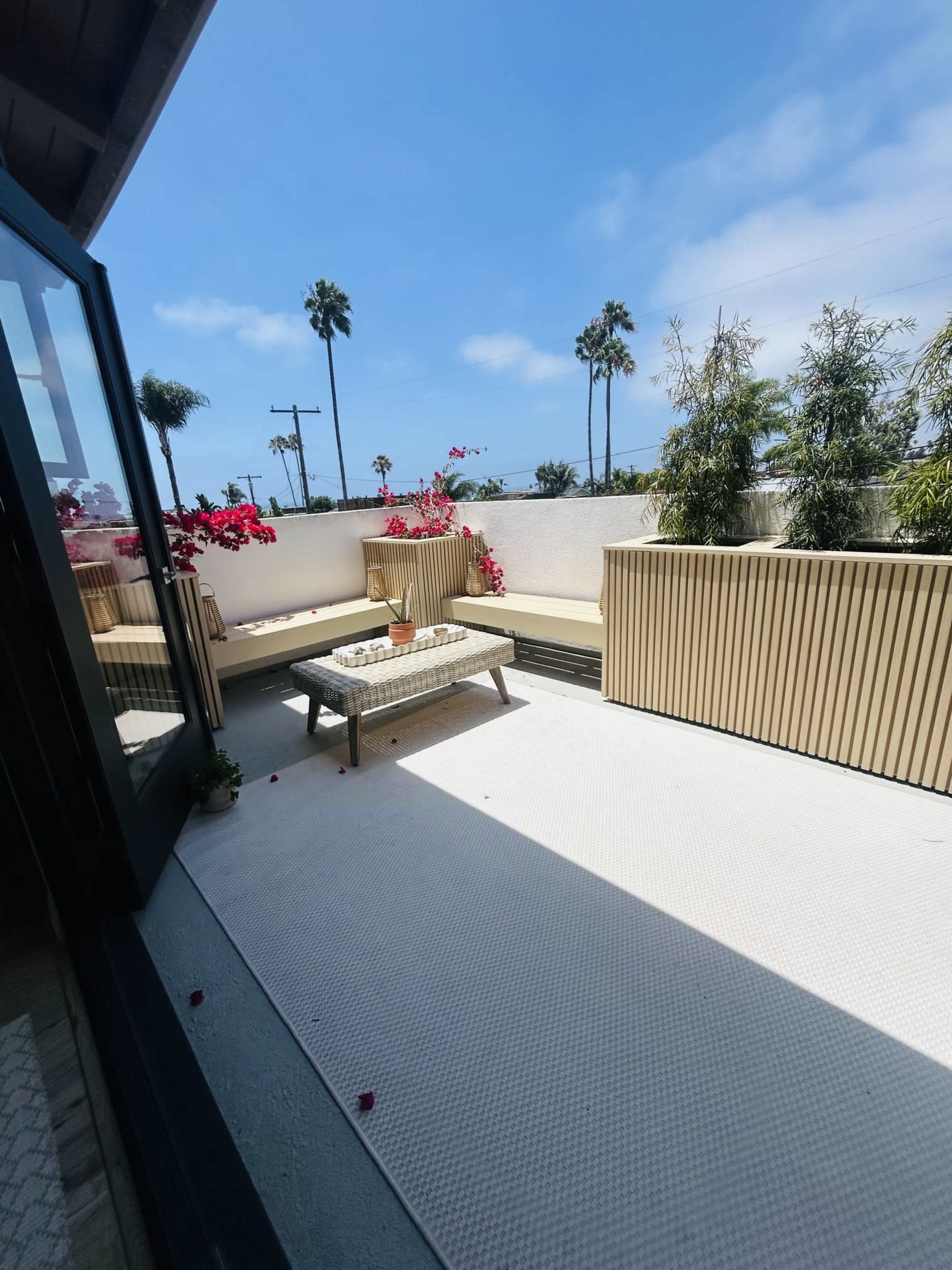The image shows a sunlit patio with wooden seating, potted plants, and a low table, framed by palm trees and blue skies.