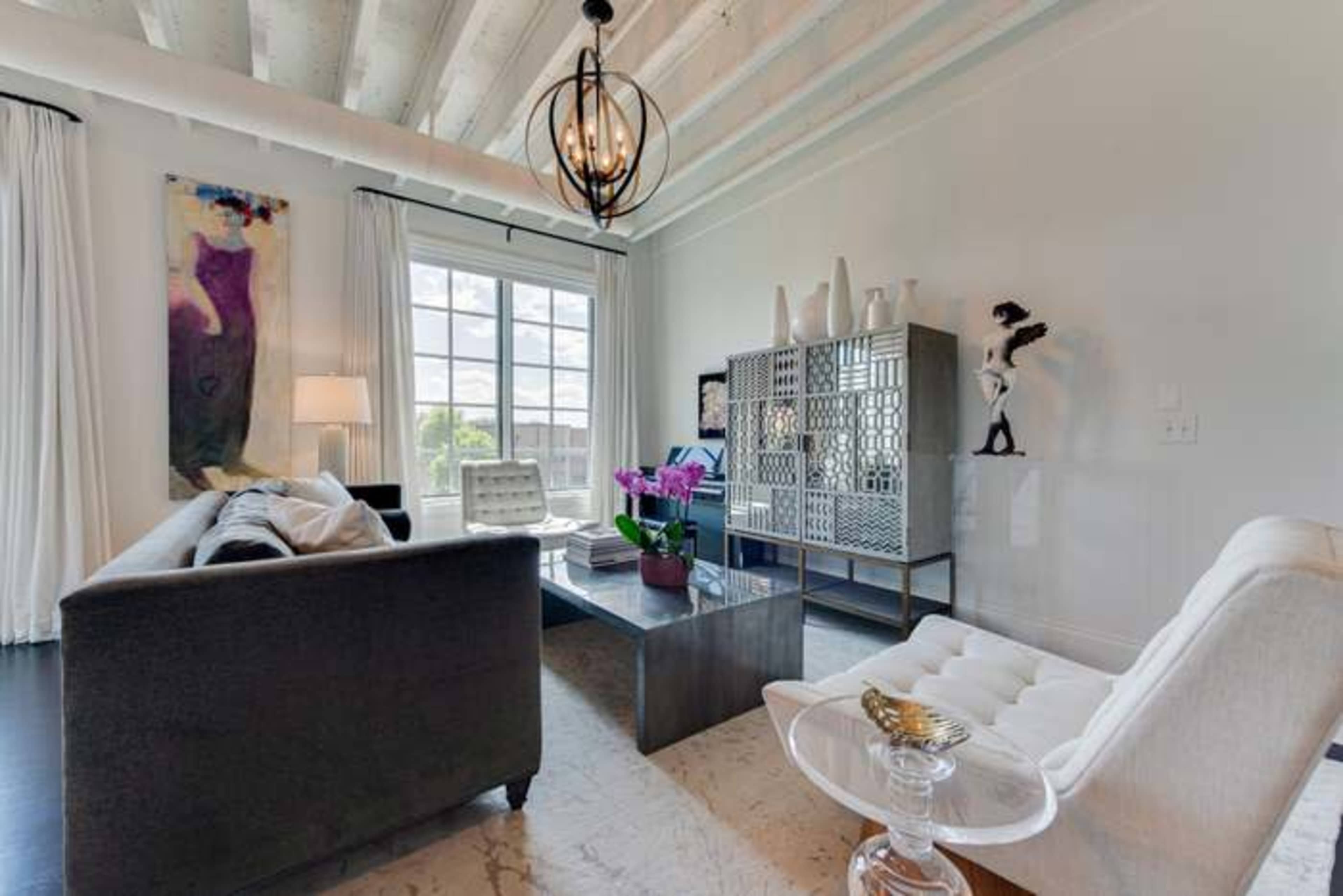 The room features a gray sofa, a glass coffee table, and a white chair, with large windows allowing natural light to enter.