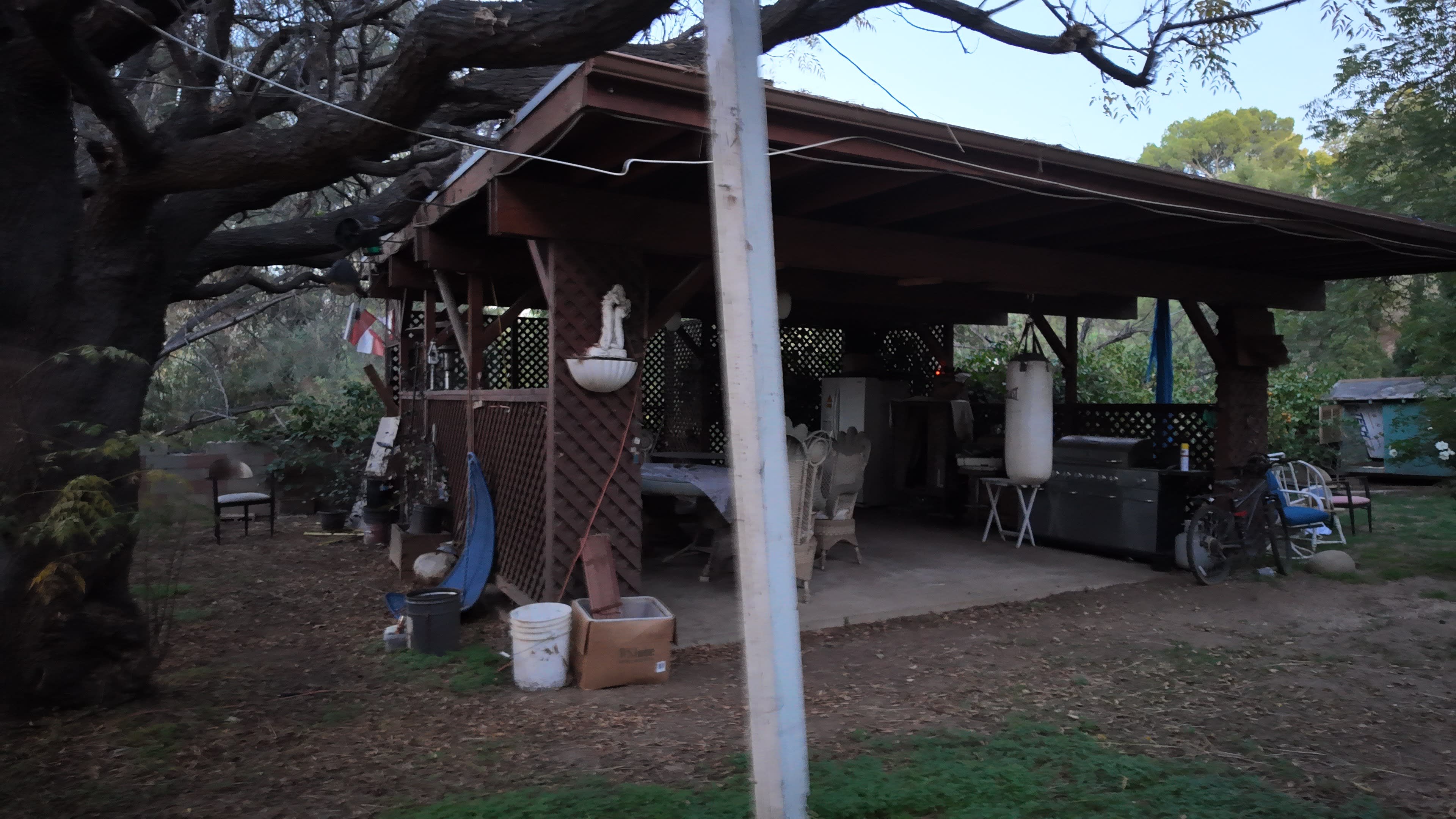 The image shows a rustic outdoor structure with a wooden frame, surrounded by trees and various items, including chairs and containers, on the ground.