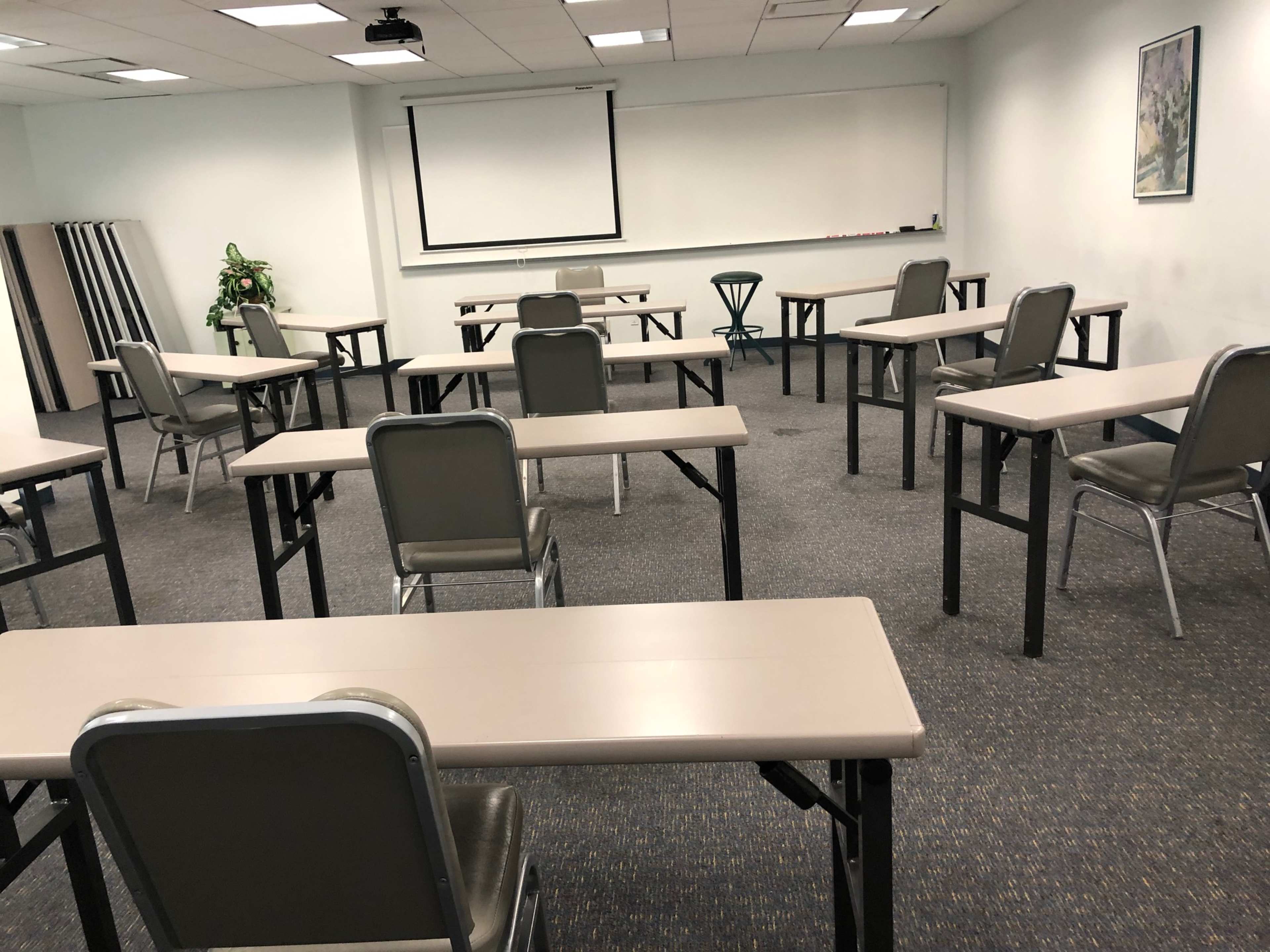 A large, empty classroom is arranged with several tables and chairs, facing a projection screen at the front.