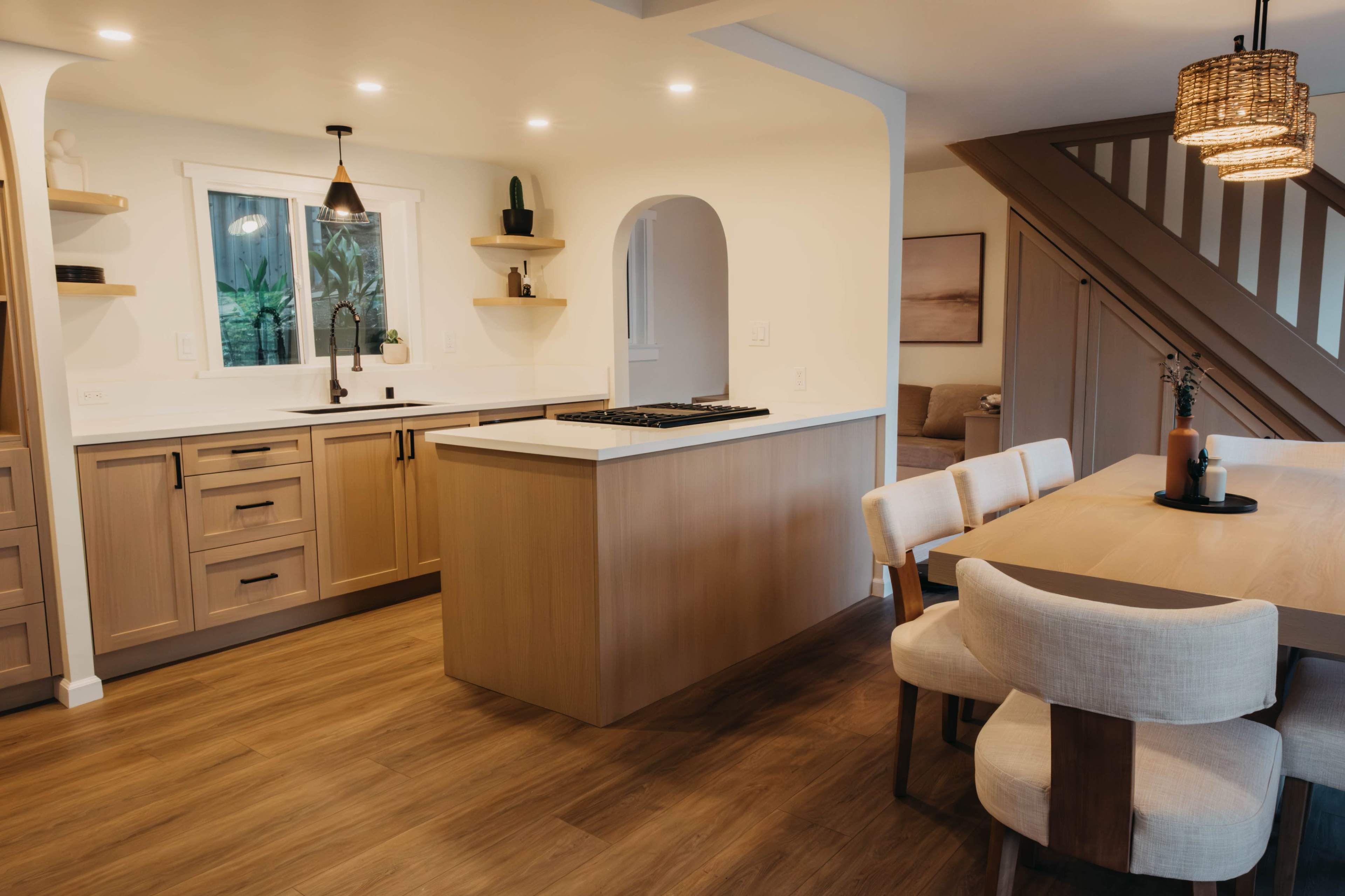 The image shows a modern kitchen with light wood cabinetry, a central island, and an adjoining dining area featuring a wooden table and upholstered chairs.