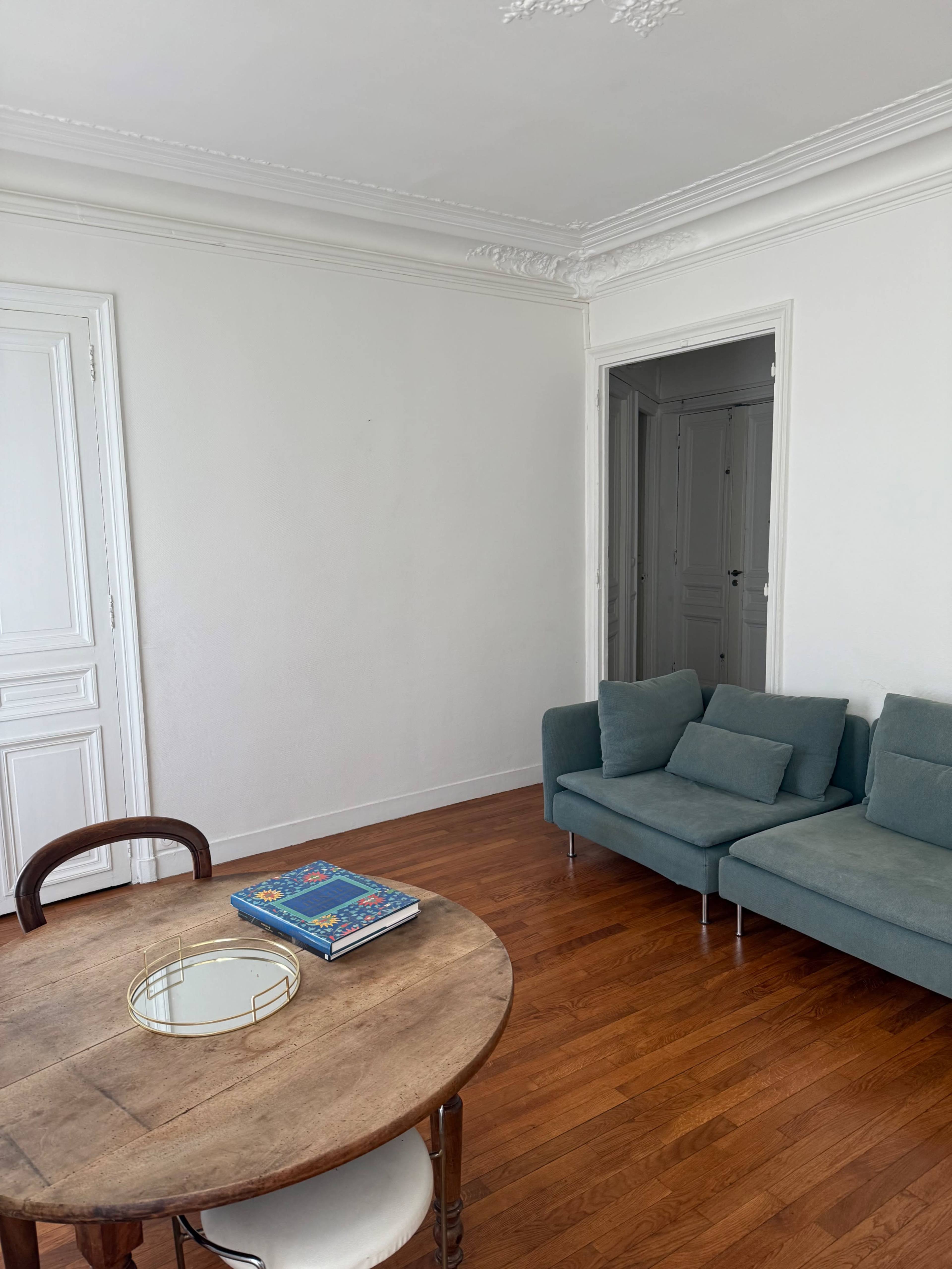The image shows a minimalist living room with a wooden table, a round mirror on it, and a teal couch against a white wall.