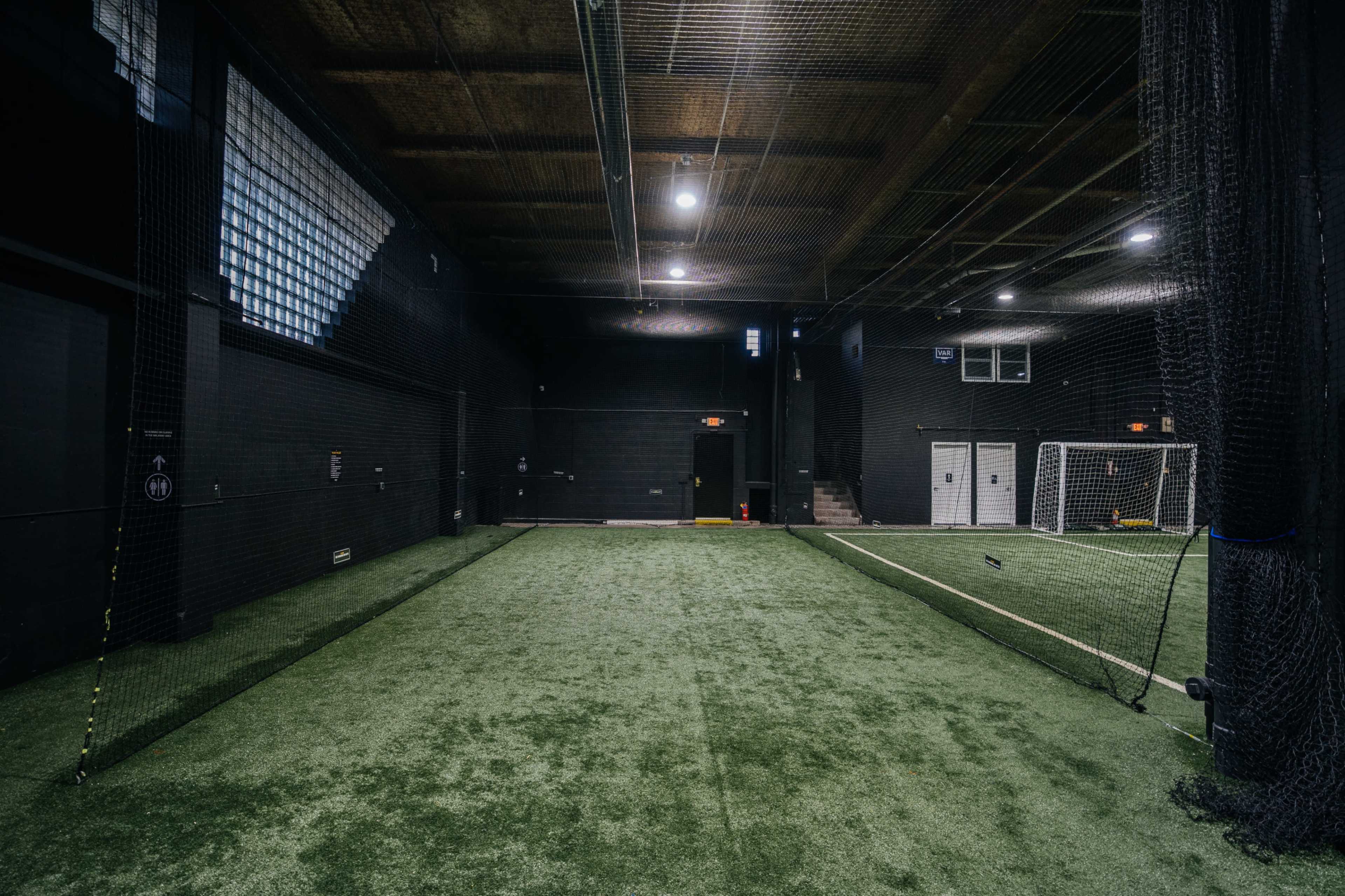 The image shows an indoor soccer training facility with artificial turf, goalposts, and netting along the sides.