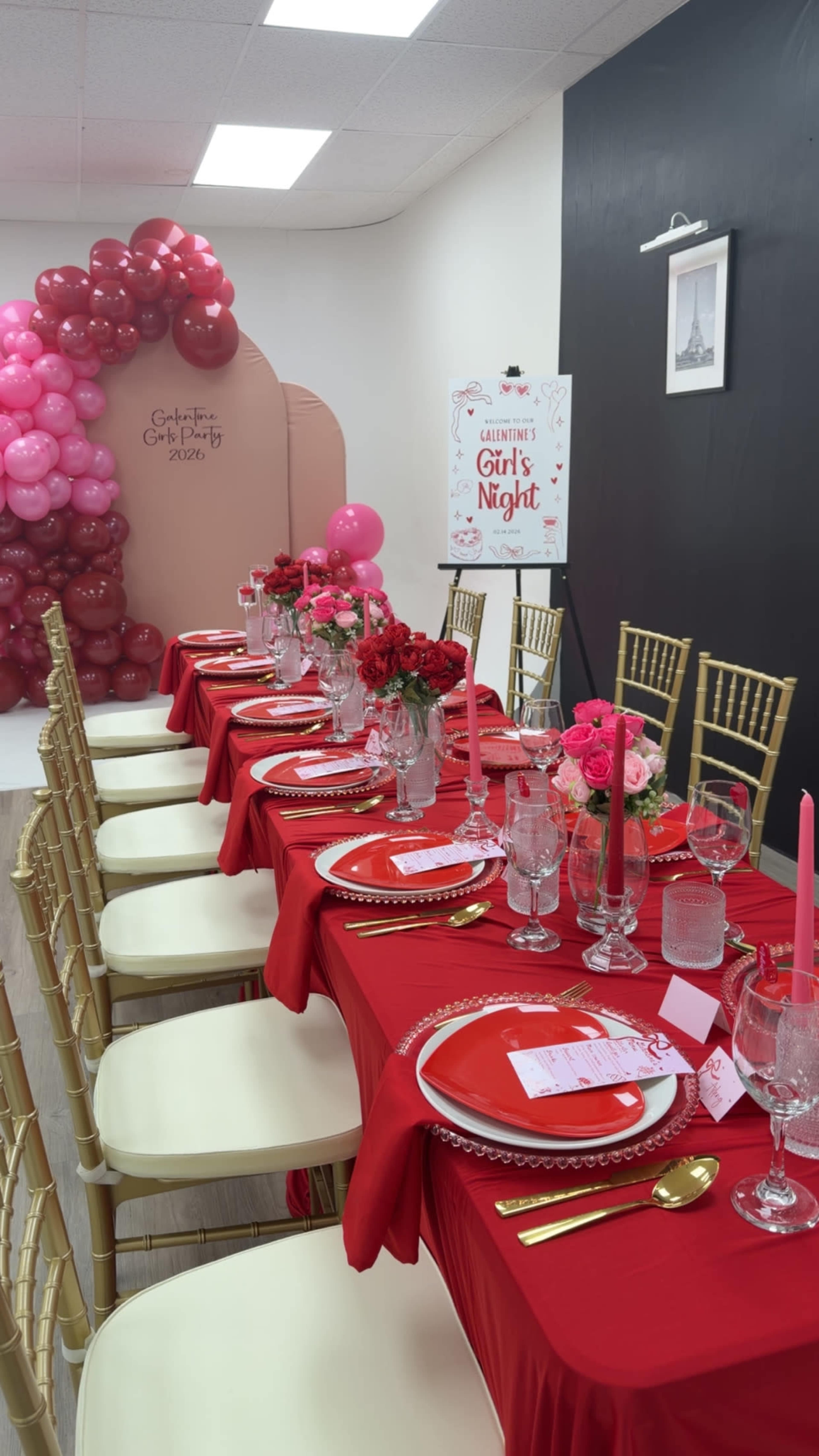 A long table is set with red tablecloths, gold utensils, and decorative centerpieces, surrounded by chairs, in a room featuring pink balloon decorations and a sign promoting a "Girl's Night" event.