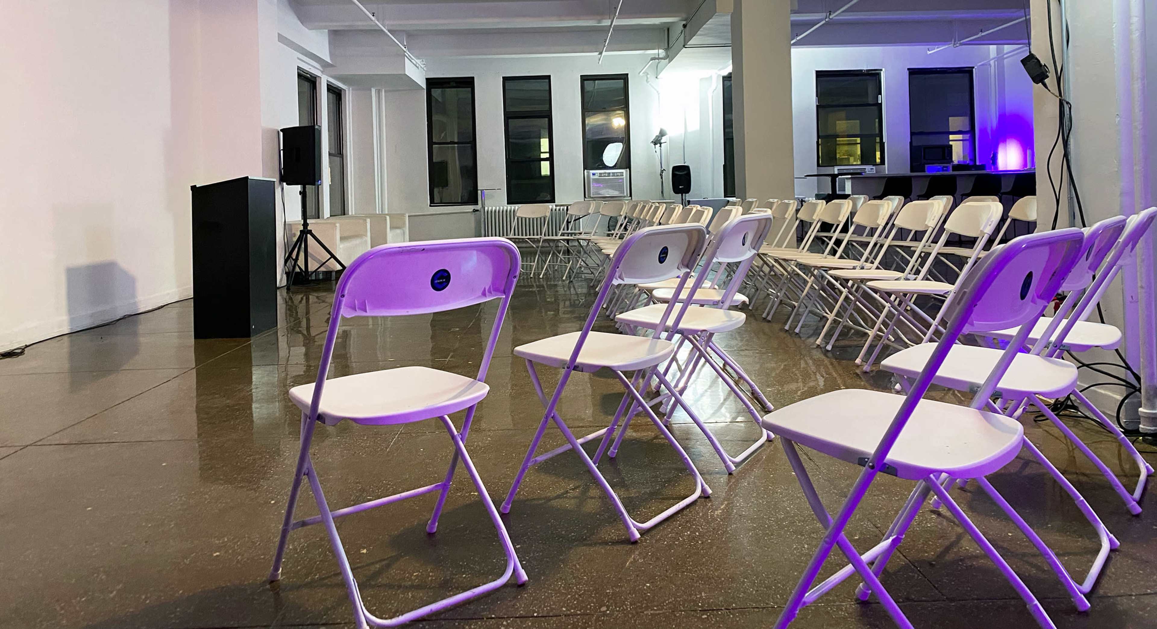 A spacious indoor venue features rows of empty white folding chairs facing a podium, with windows allowing natural light and ambient purple lighting.