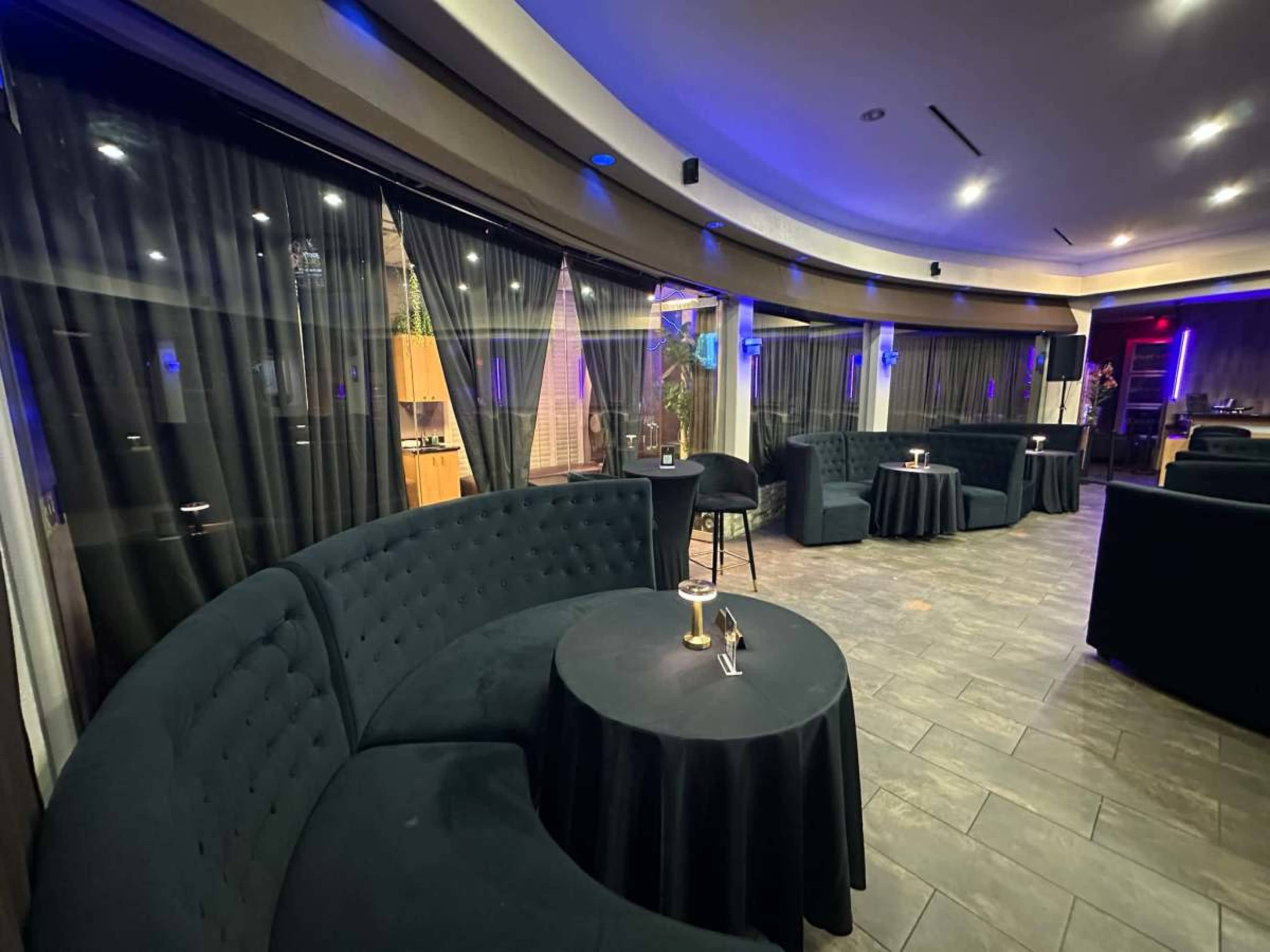 The image shows a dimly lit lounge area featuring curved velvet seating, small tables, and large windows with sheer black curtains.