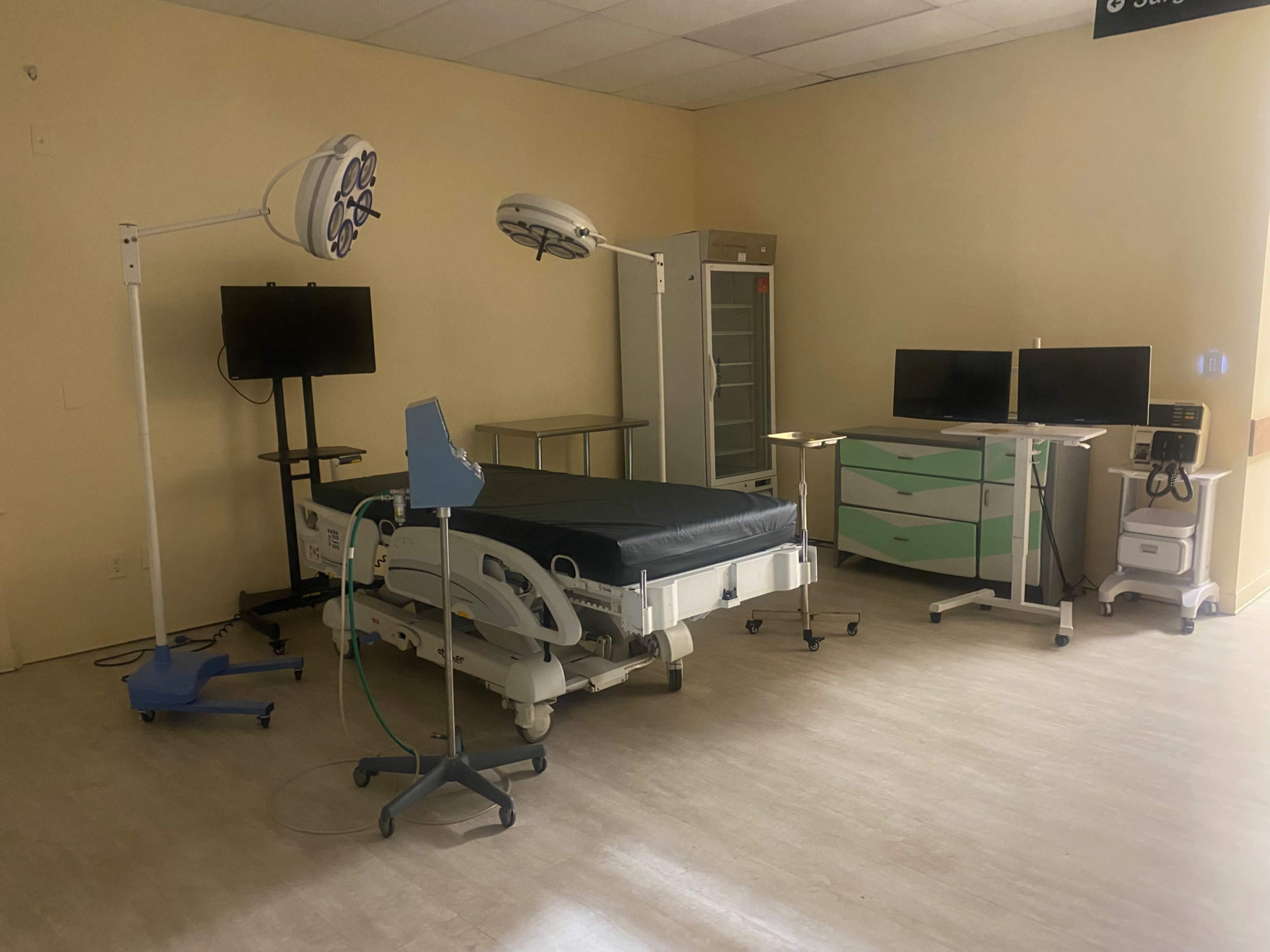 The image shows a hospital room equipped with a surgical bed, two monitors, a medical cart, and a storage cabinet.