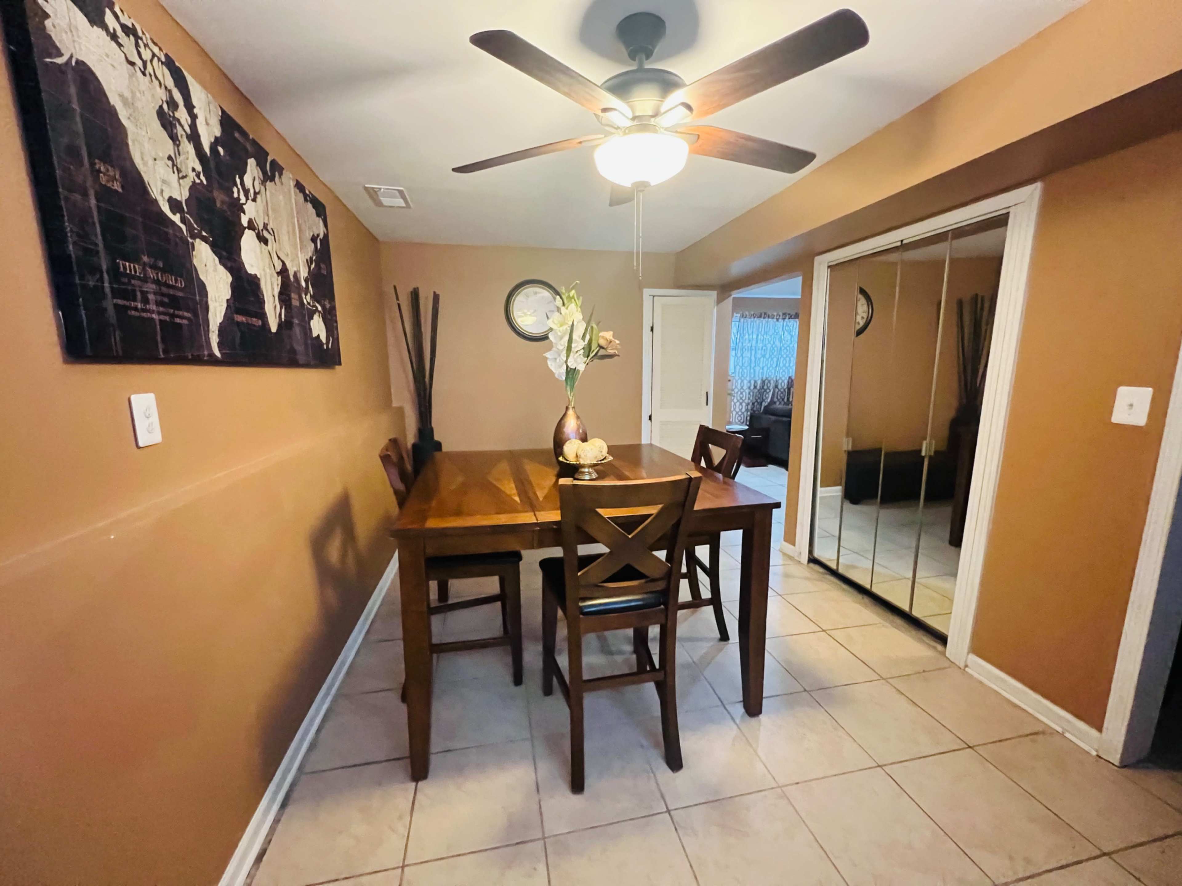 A dining area features a wooden table with four chairs, a ceiling fan, and a wall adorned with a world map, alongside a mirrored closet.