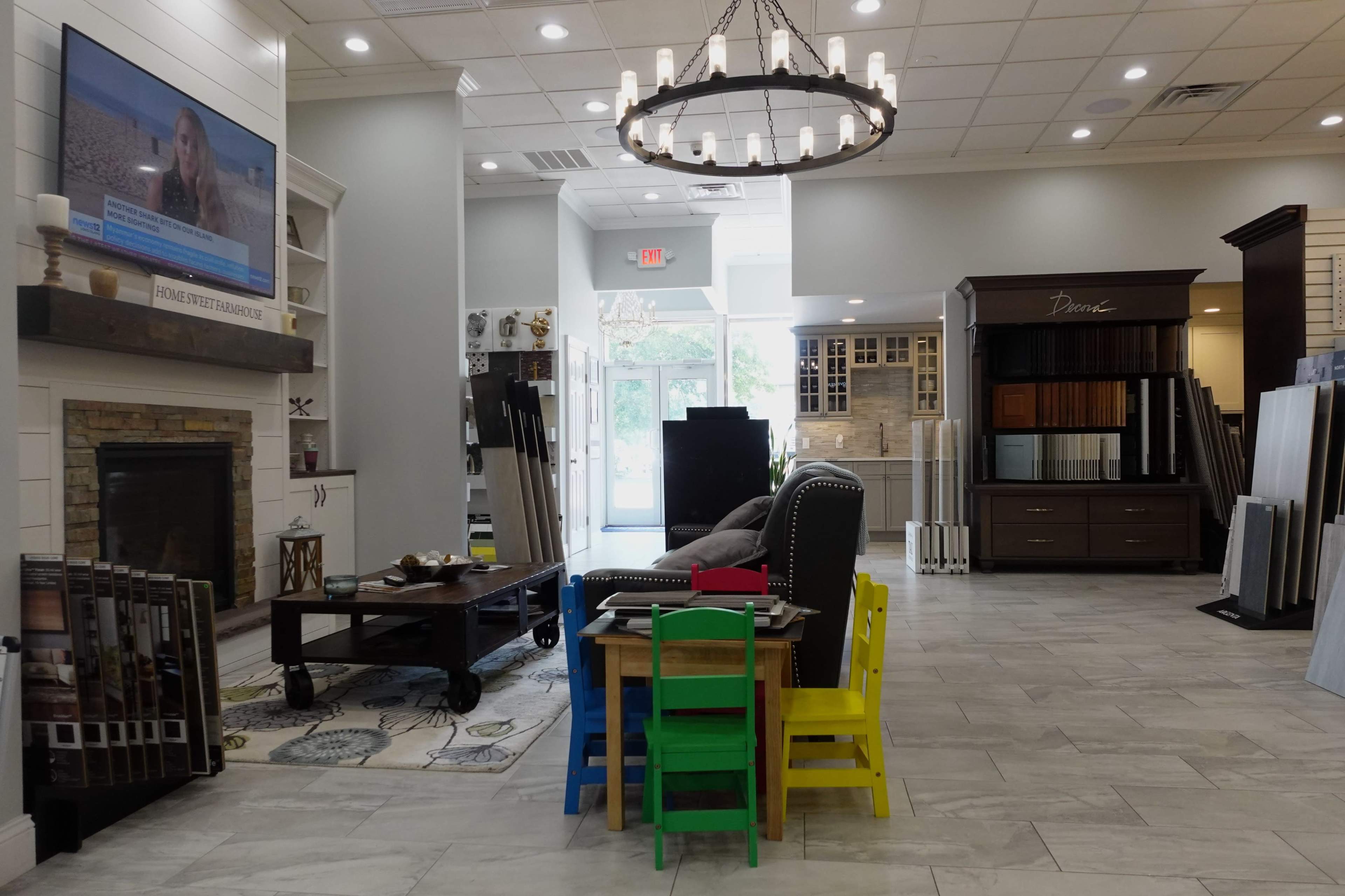 A showroom featuring a fireplace display, a television, a seating area with colorful children's chairs, and various samples of flooring and cabinetry on the walls.
