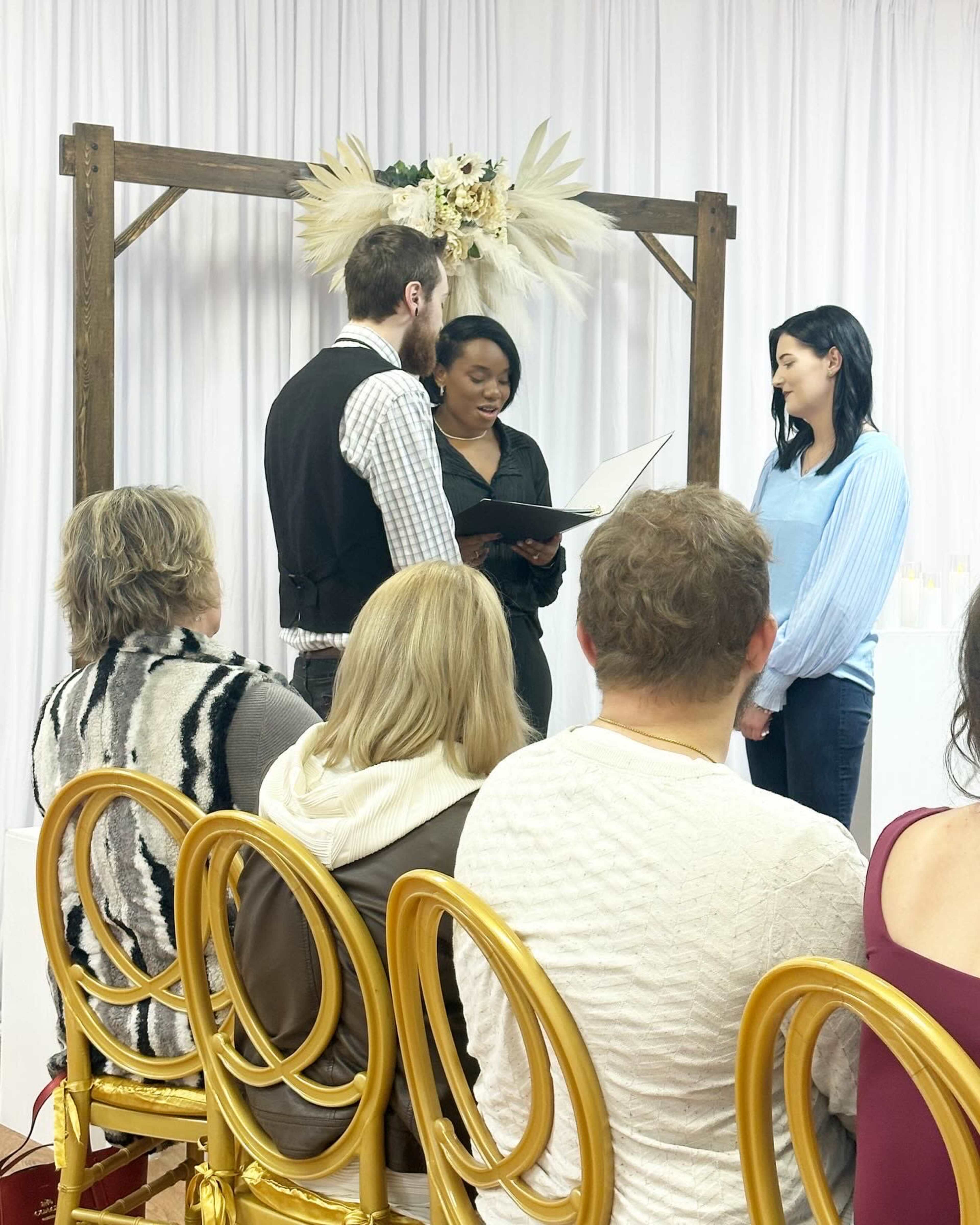 A couple stands in front of a person reading from a booklet during a ceremony, with guests seated on gold chairs in the foreground.
