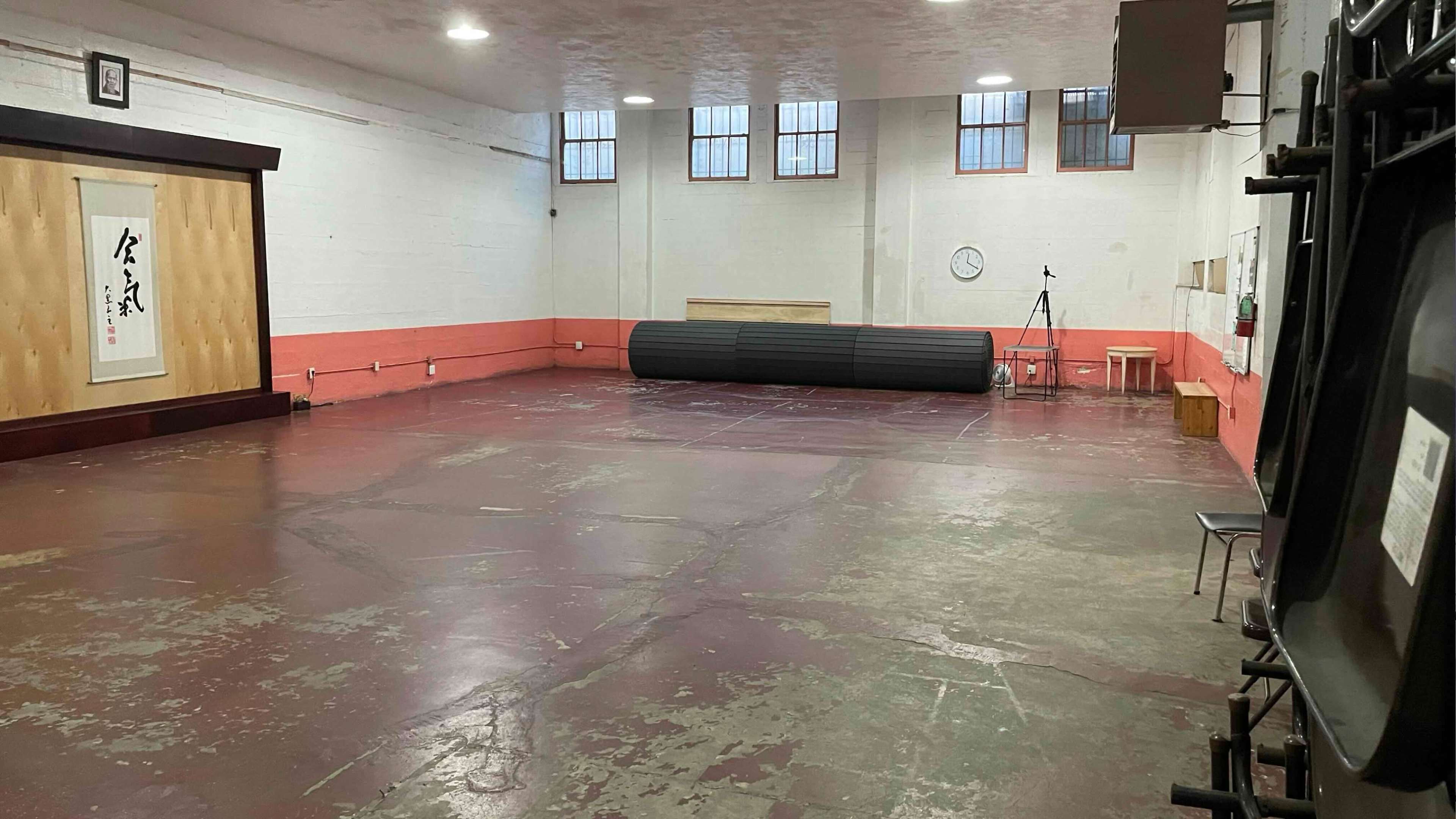 The image shows an empty gymnasium with a polished concrete floor, dim lighting, and a large black mat rolled up against the wall.