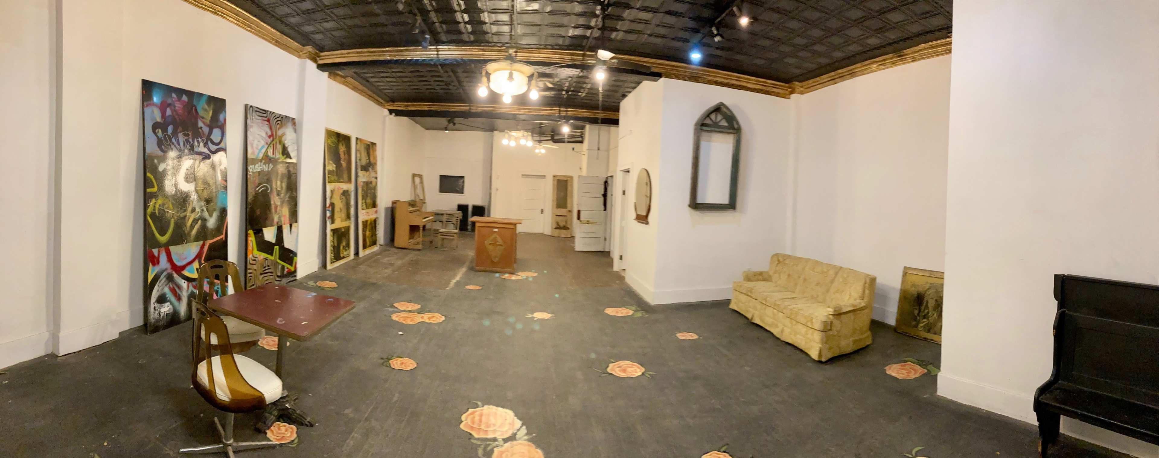 A spacious, well-lit room with a sofa, a table and chair, several large paintings on the wall, and floral-patterned flooring.