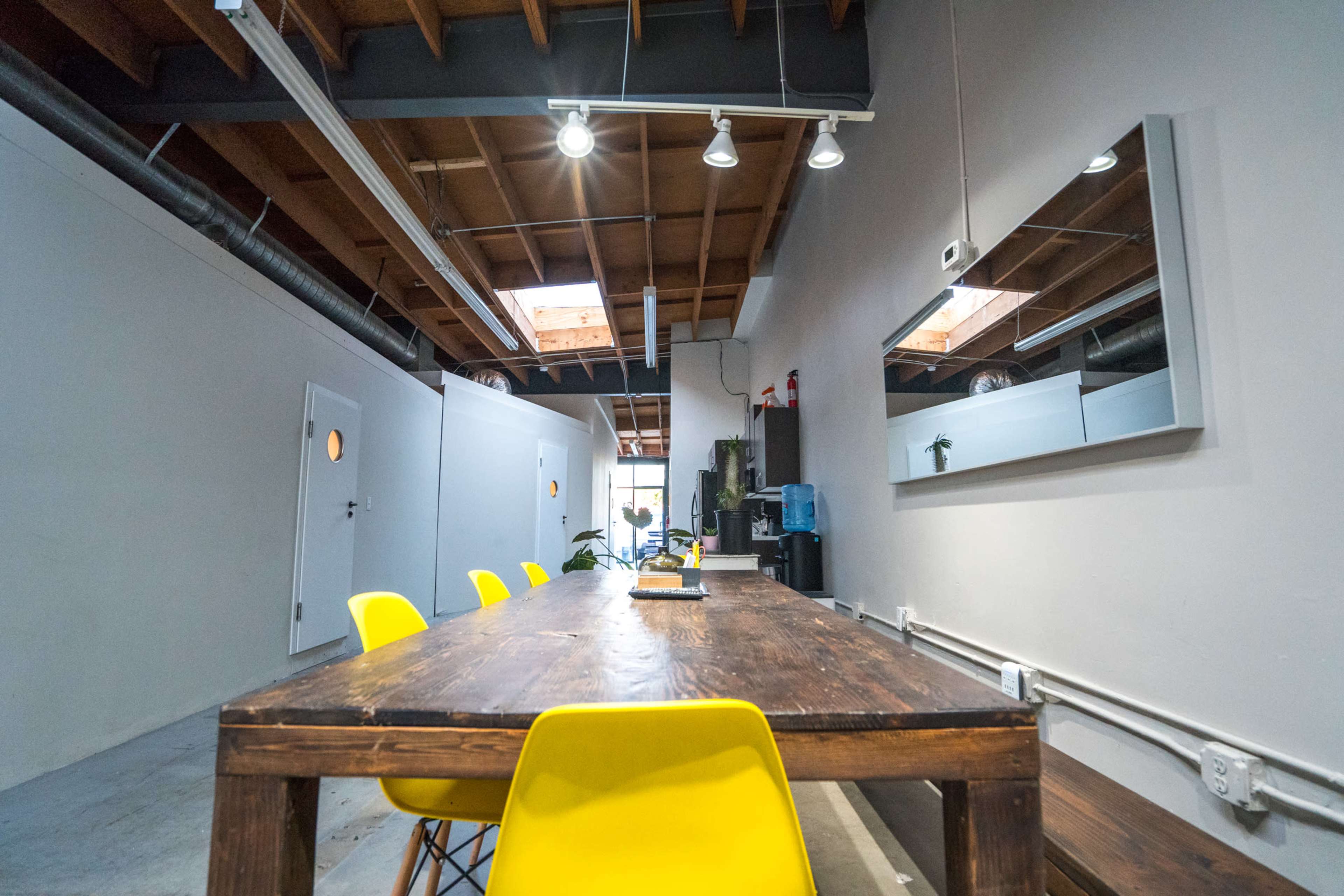 A long wooden table with yellow chairs in a brightly lit, industrial-style workspace featuring a mirror and a water cooler along the walls.
