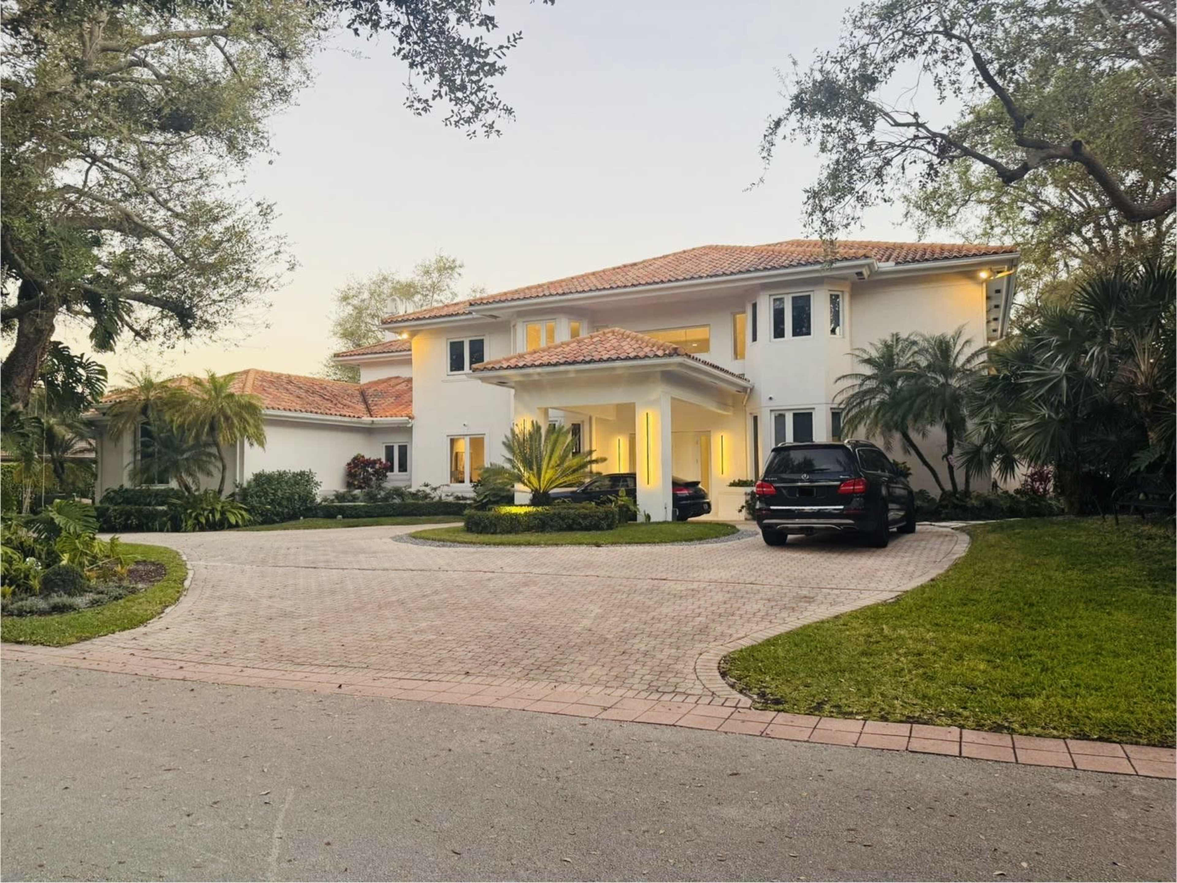 A large, two-story house with a tile roof is surrounded by lush landscaping and features a circular driveway with two parked cars.