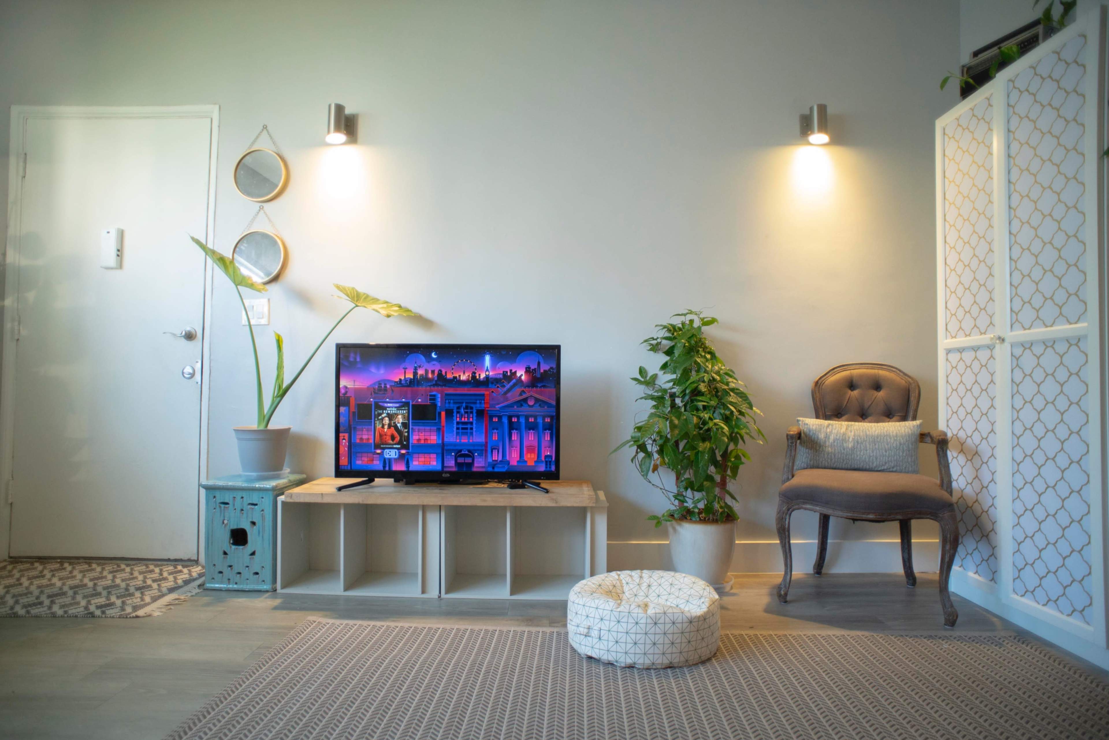 A living room features a television on a wooden stand, surrounded by plants, a decorative chair, and wall-mounted lights.