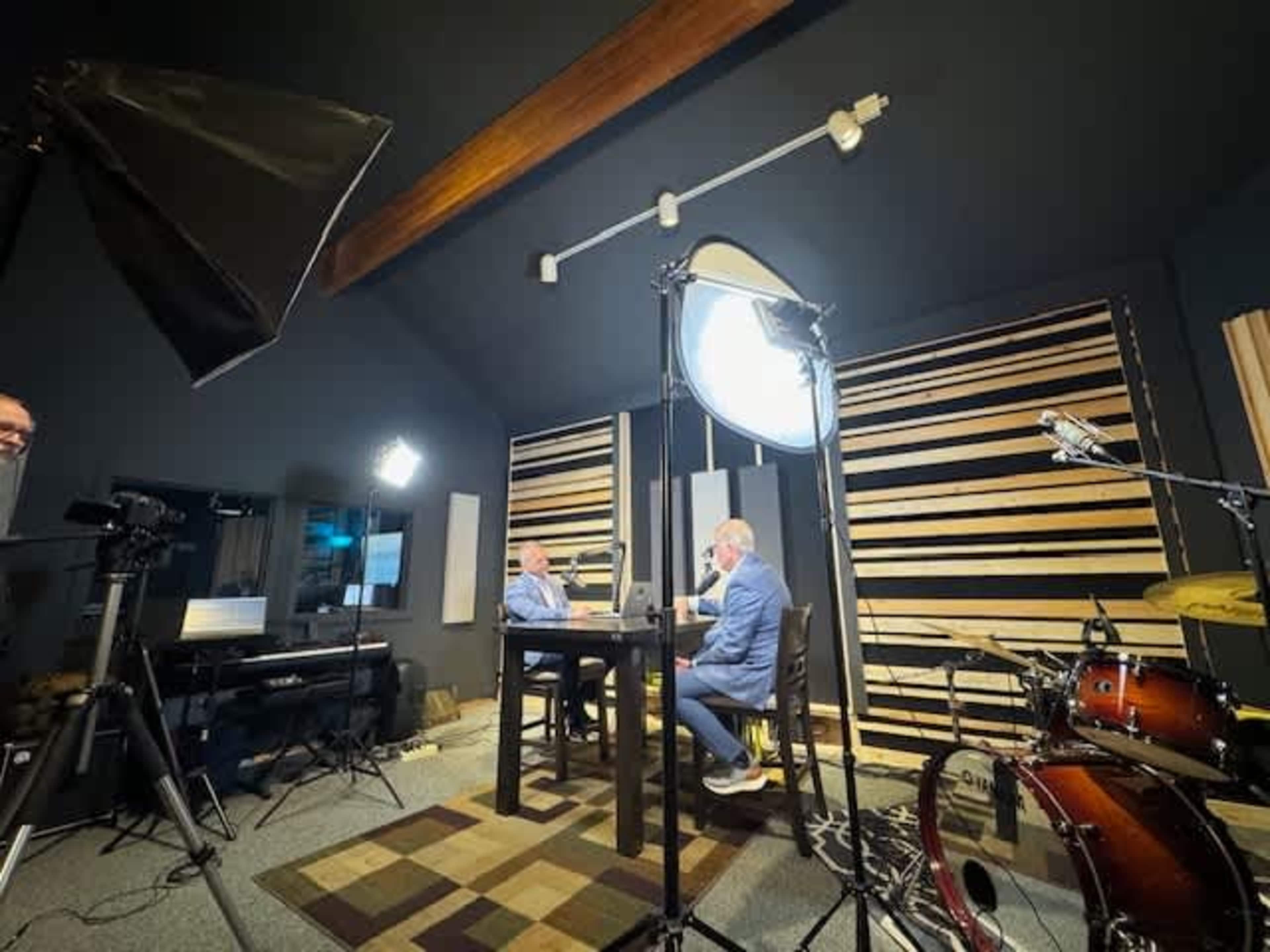 Two men are sitting at a table in a recording studio, surrounded by professional lighting and audio equipment, with a drum set visible in the corner.
