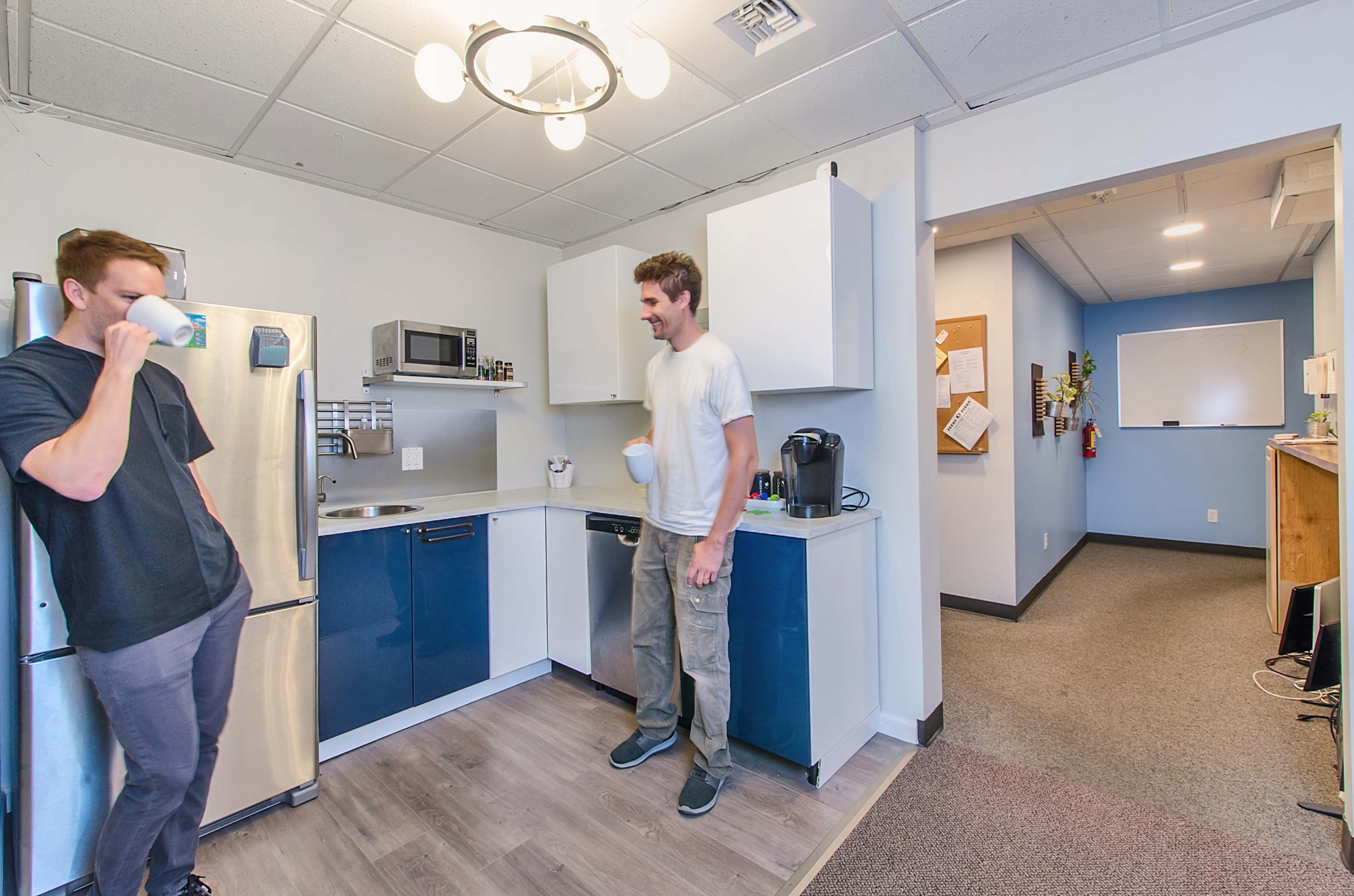 Two men are standing in a kitchen area with a refrigerator, microwave, and coffee maker while one drinks from a cup and the other holds a mug.