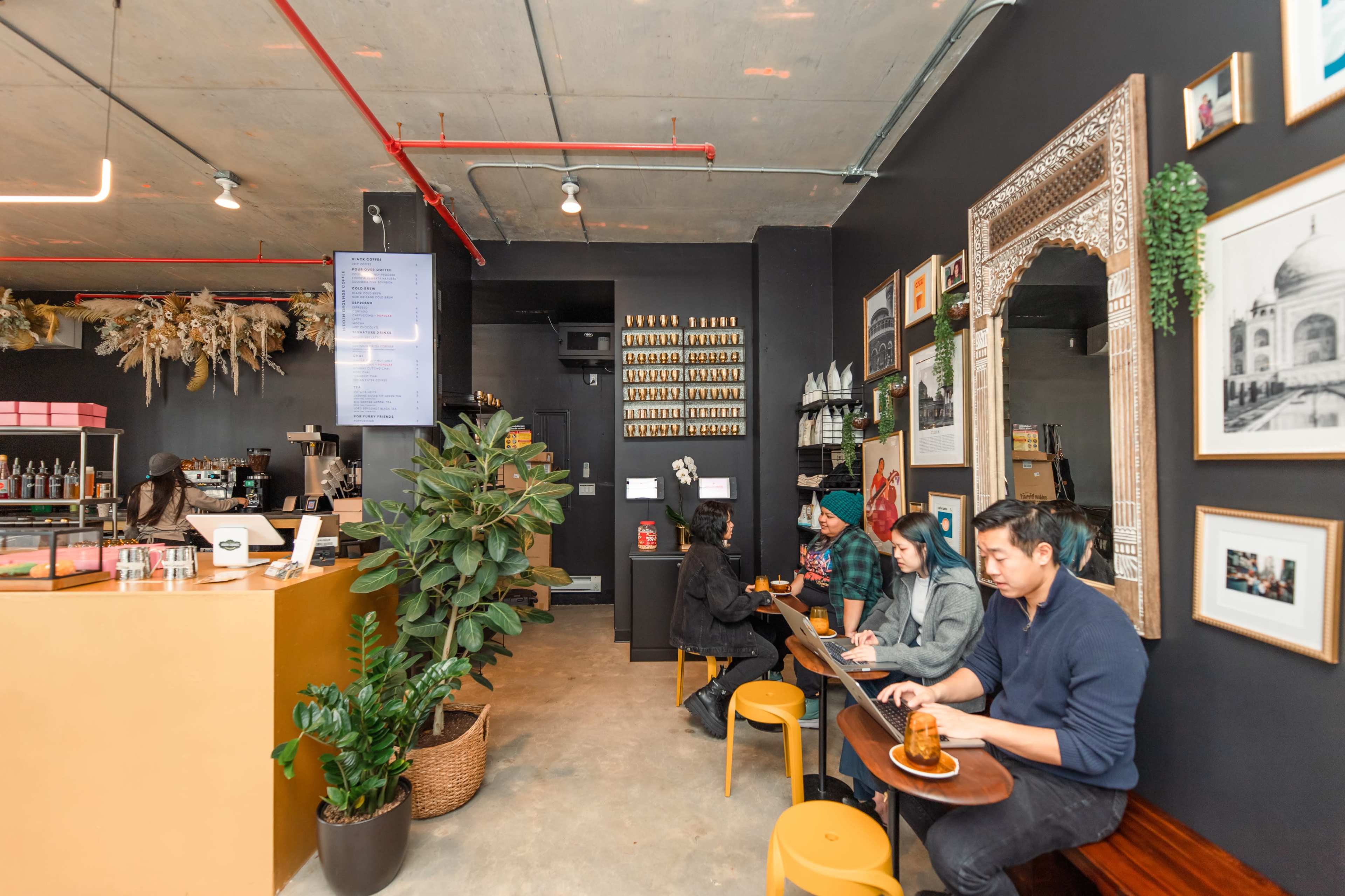 A modern café interior with customers sitting at tables working on laptops, surrounded by plants and decorative art on the walls.
