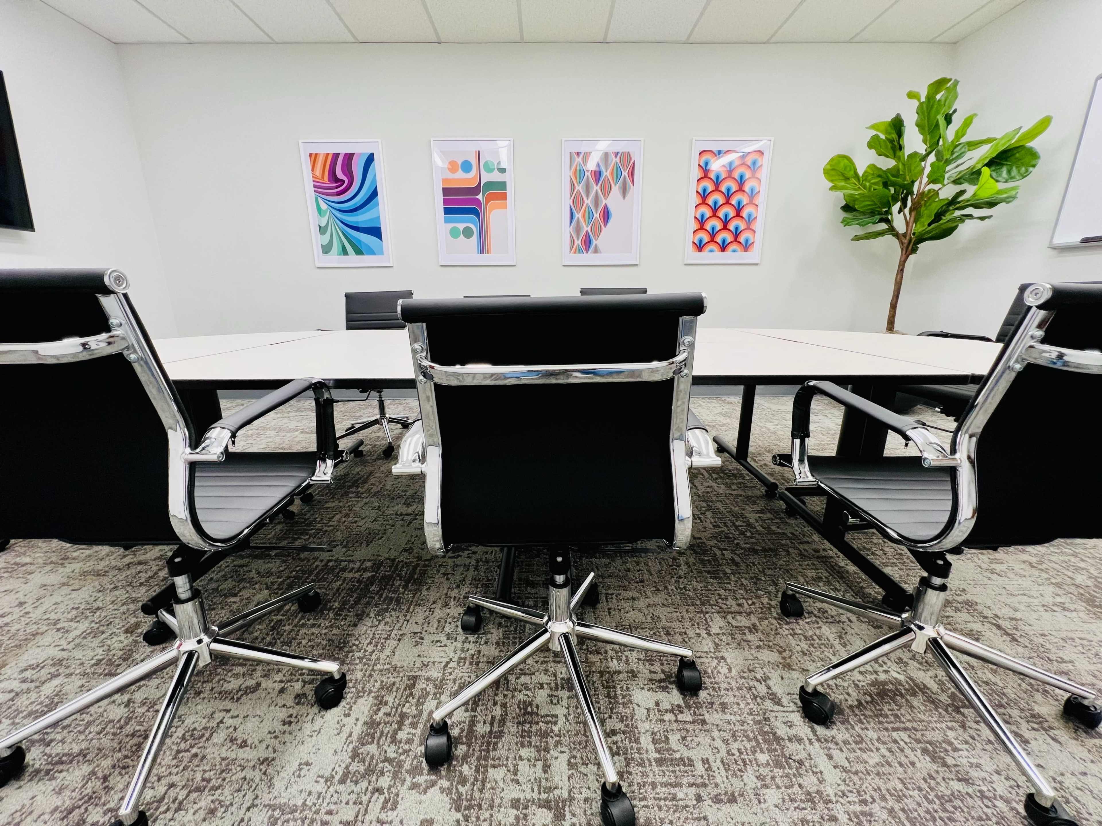 A conference room with a large, rectangular table surrounded by black office chairs, featuring colorful framed artwork on the walls and a decorative plant in one corner.