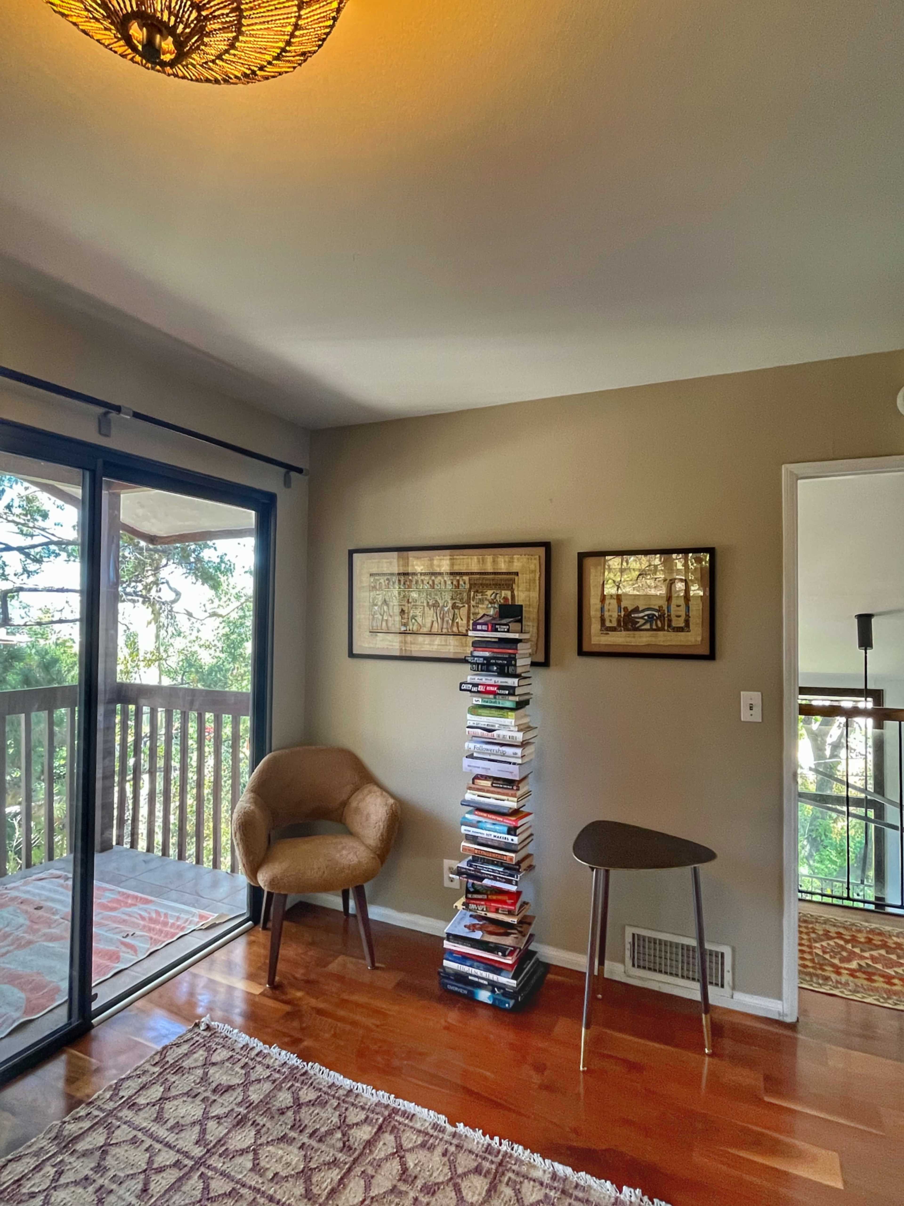 The image shows a corner of a room with a chair near a stack of books on a small table, framed artwork on the wall, and large glass doors leading to a balcony.