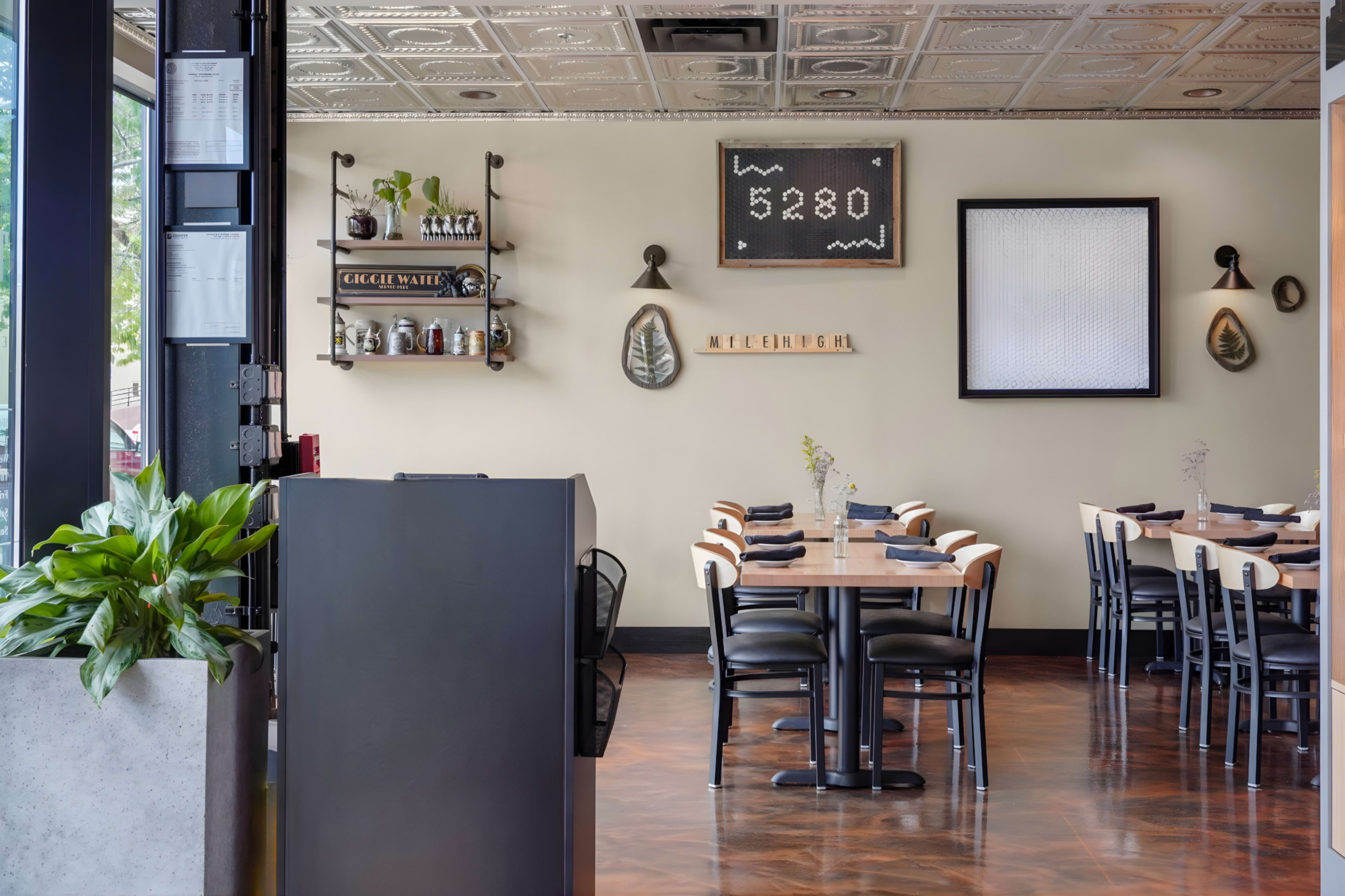 The image displays a modern restaurant interior featuring wooden tables and black chairs, with plants and wall decor including a clock and signage in a well-lit space.