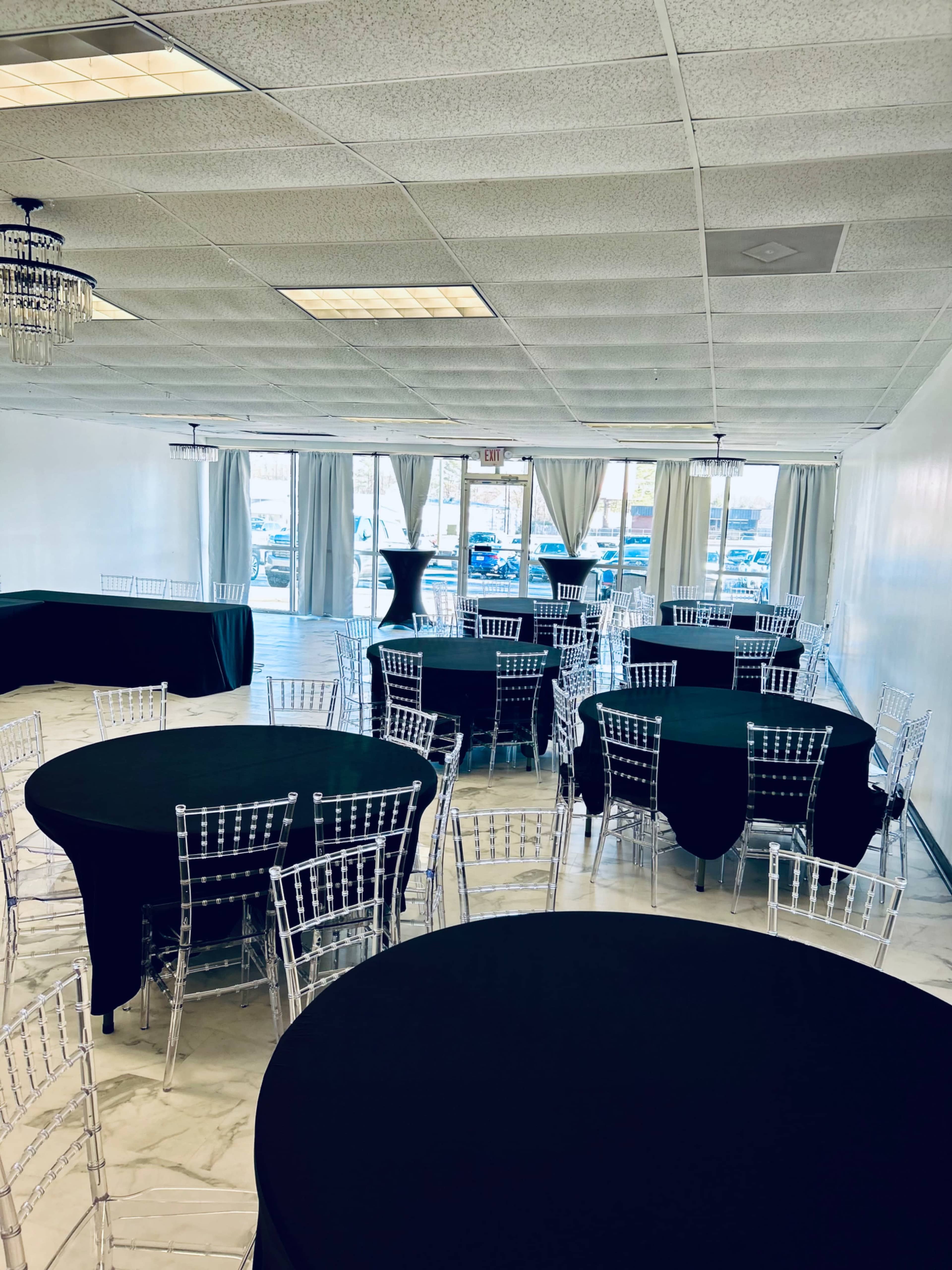 The image shows a spacious event room with multiple round tables covered in black tablecloths and complemented by clear chiavari chairs, along with large windows draped with curtains.