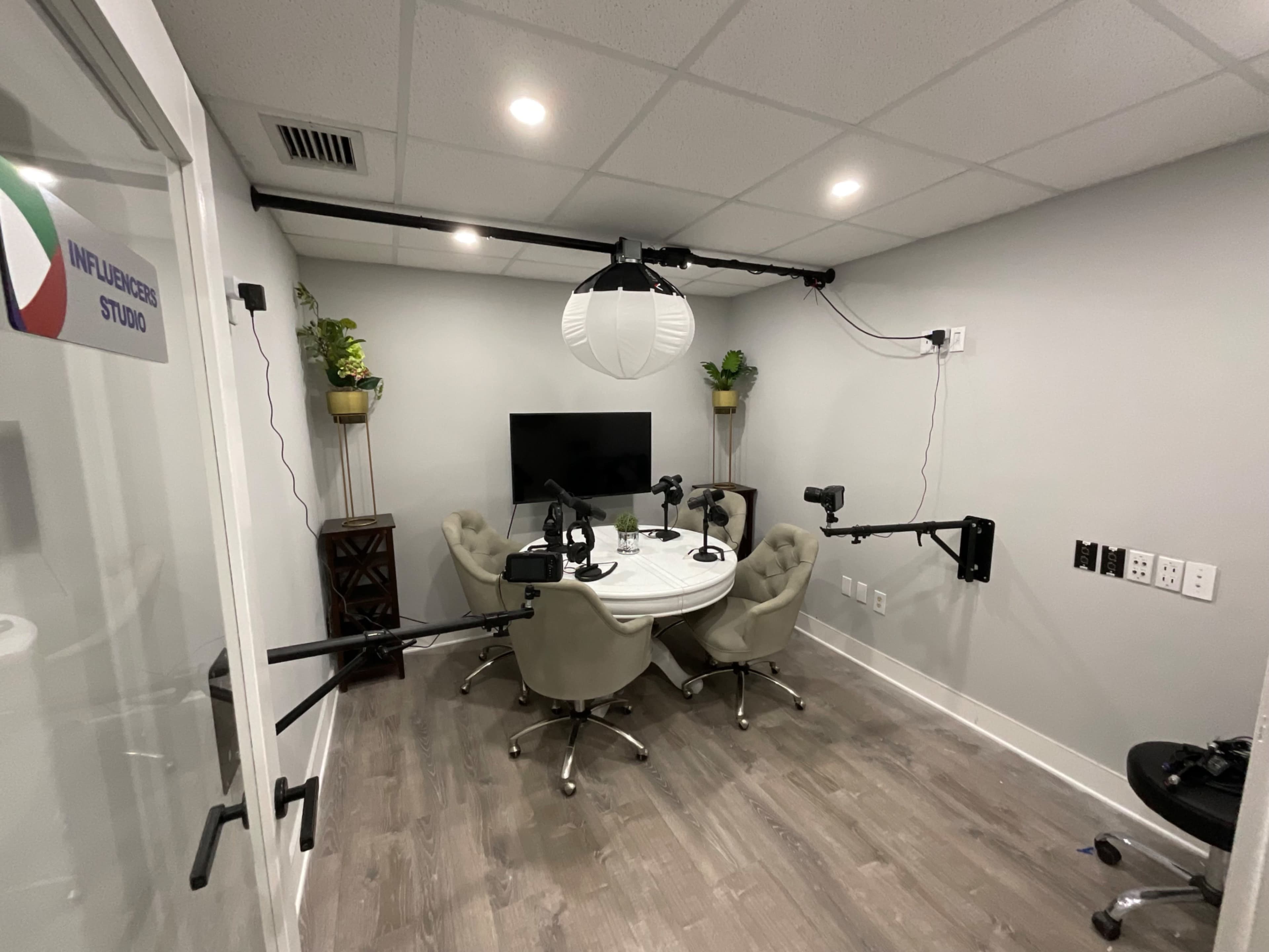 A small recording studio with a round table surrounded by four chairs, equipped with microphones and cameras, and featuring a television on the wall.