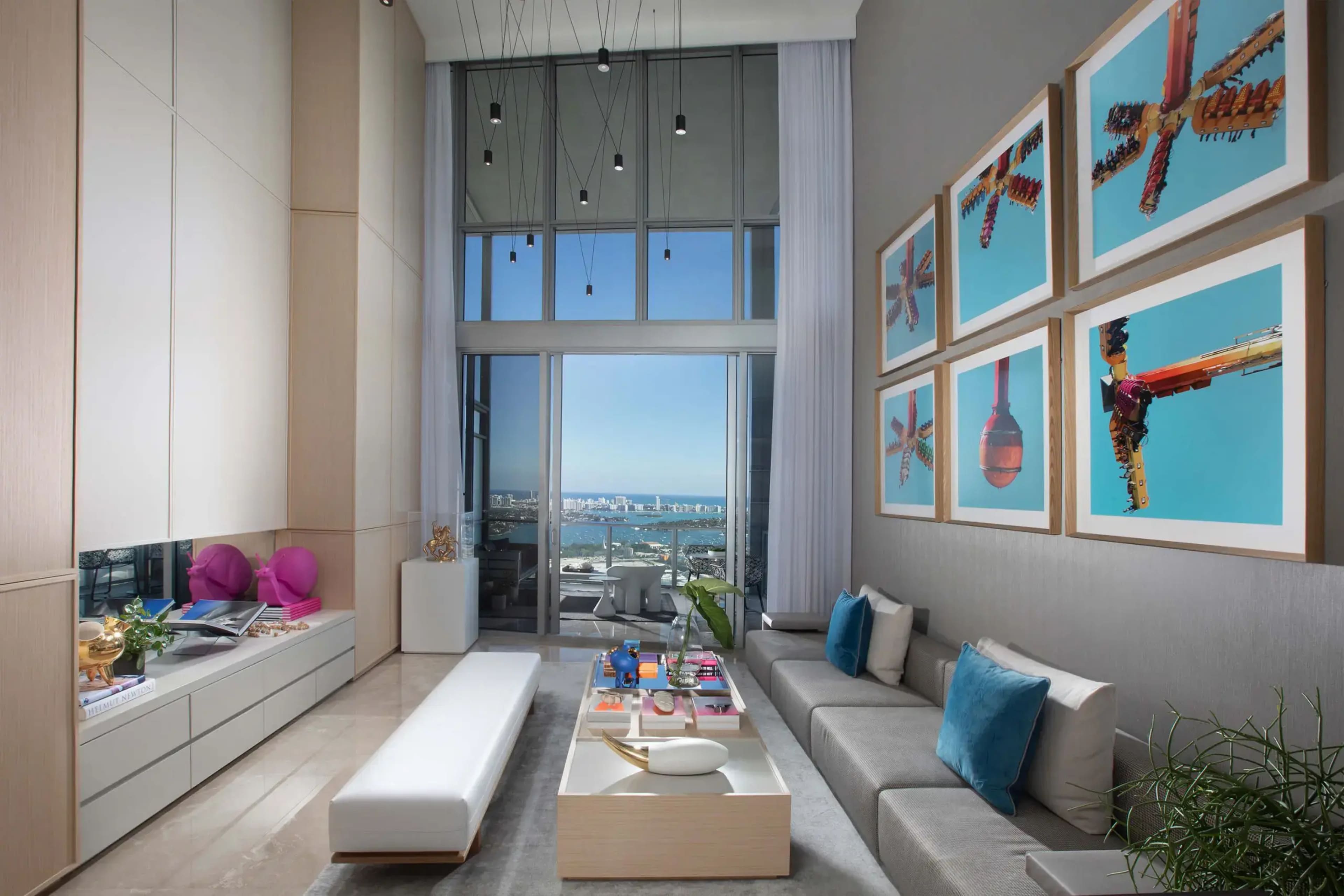 A modern living room with large windows overlooking a cityscape, featuring a long sofa, an elegant coffee table, and colorful artwork on the walls.