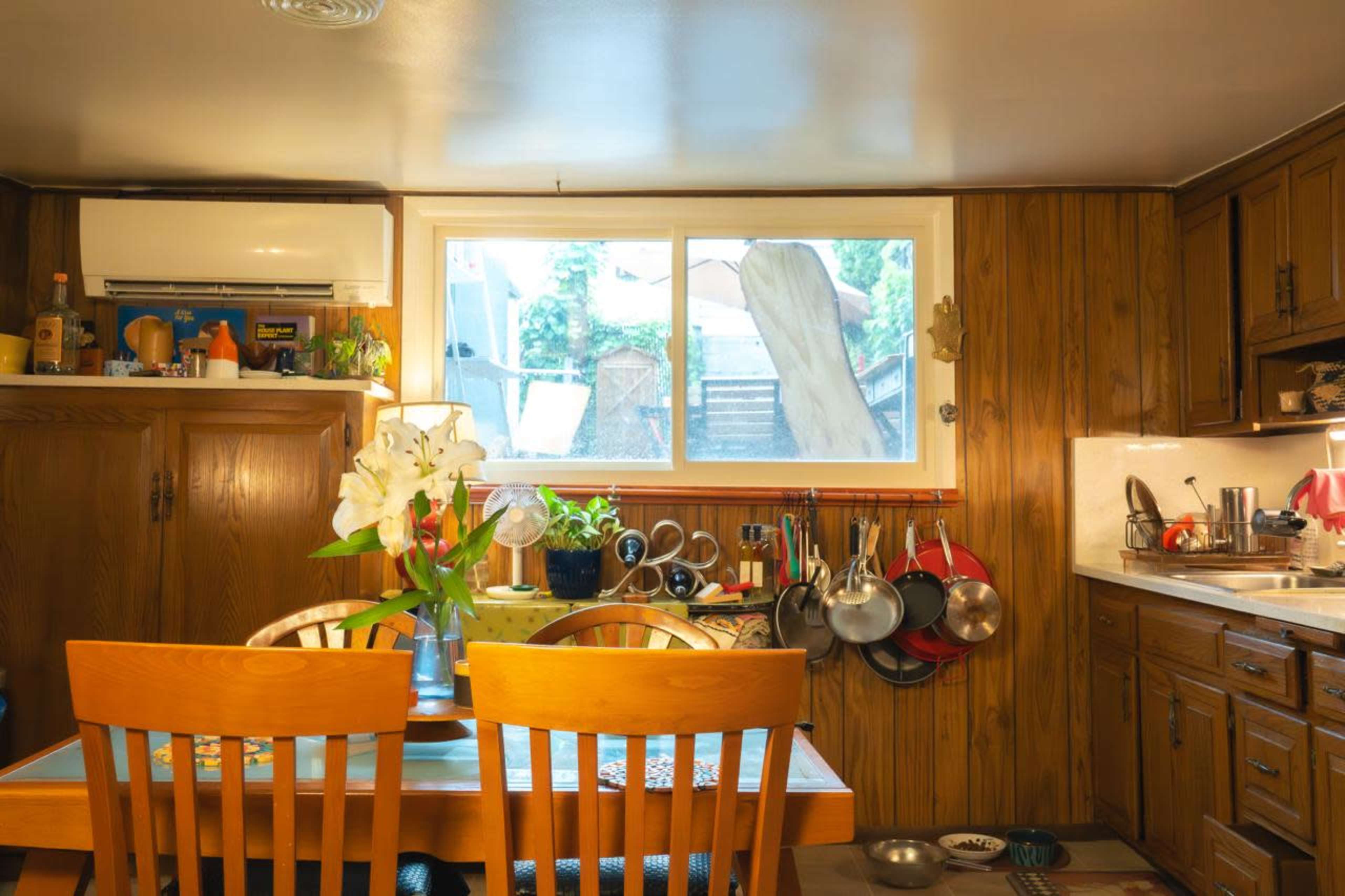 A small kitchen features a wooden dining table, chairs, and various cooking utensils hanging on the wall, with a window overlooking an outdoor area.