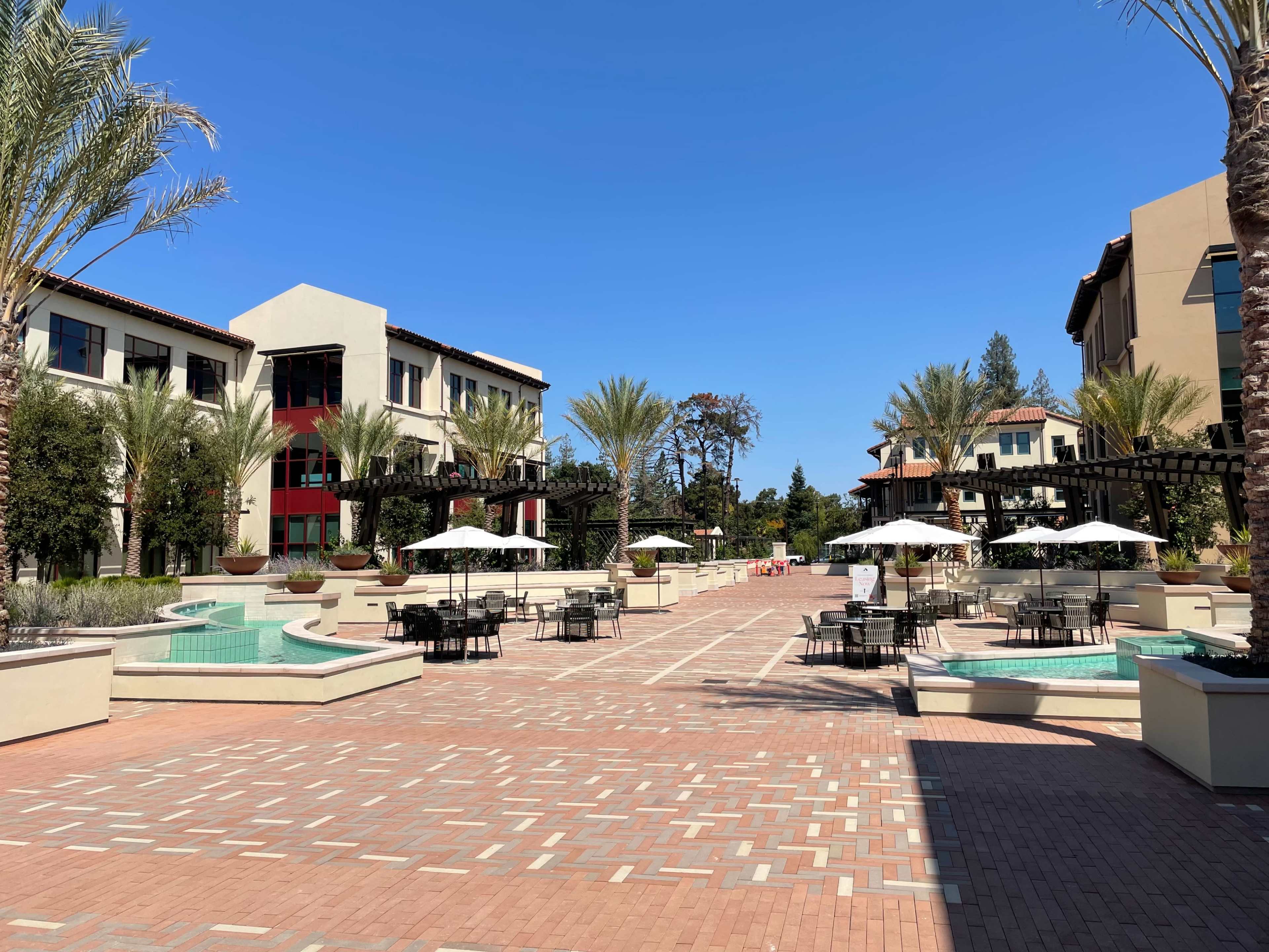 A spacious courtyard lined with palm trees, featuring seating areas and decorative fountains between modern buildings.