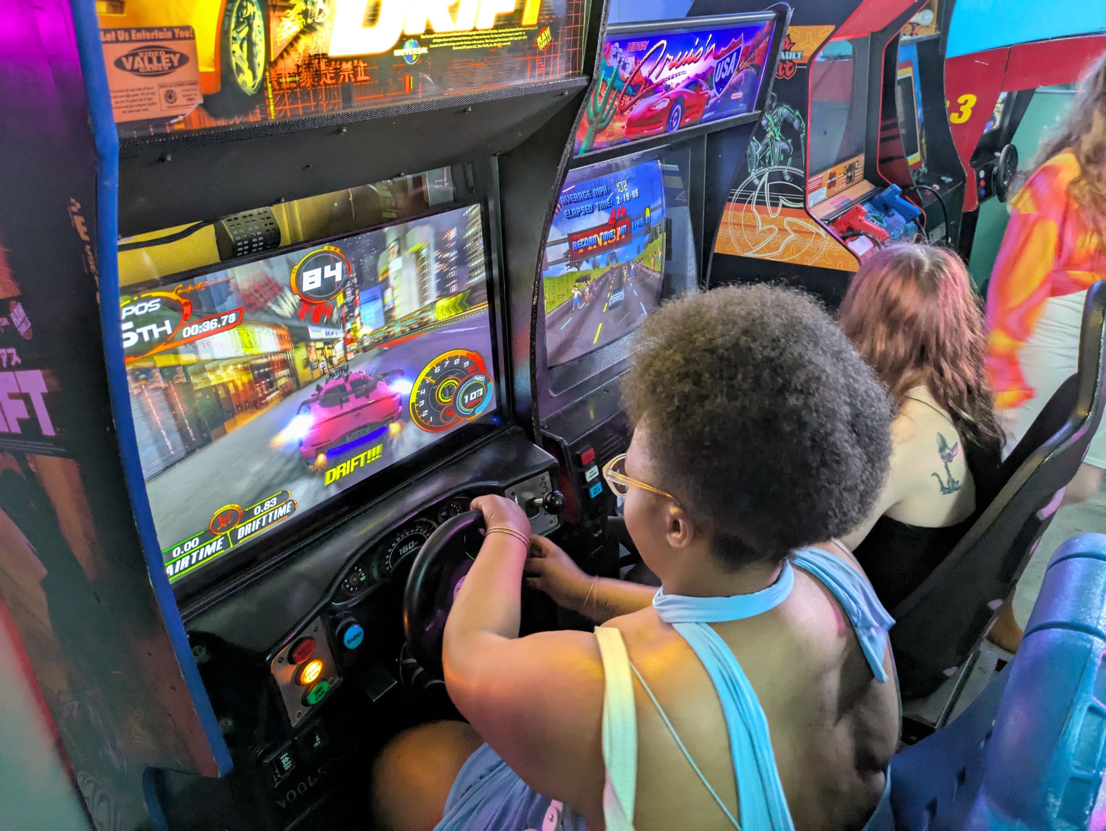 Two people are playing racing arcade games in a bright arcade setting.