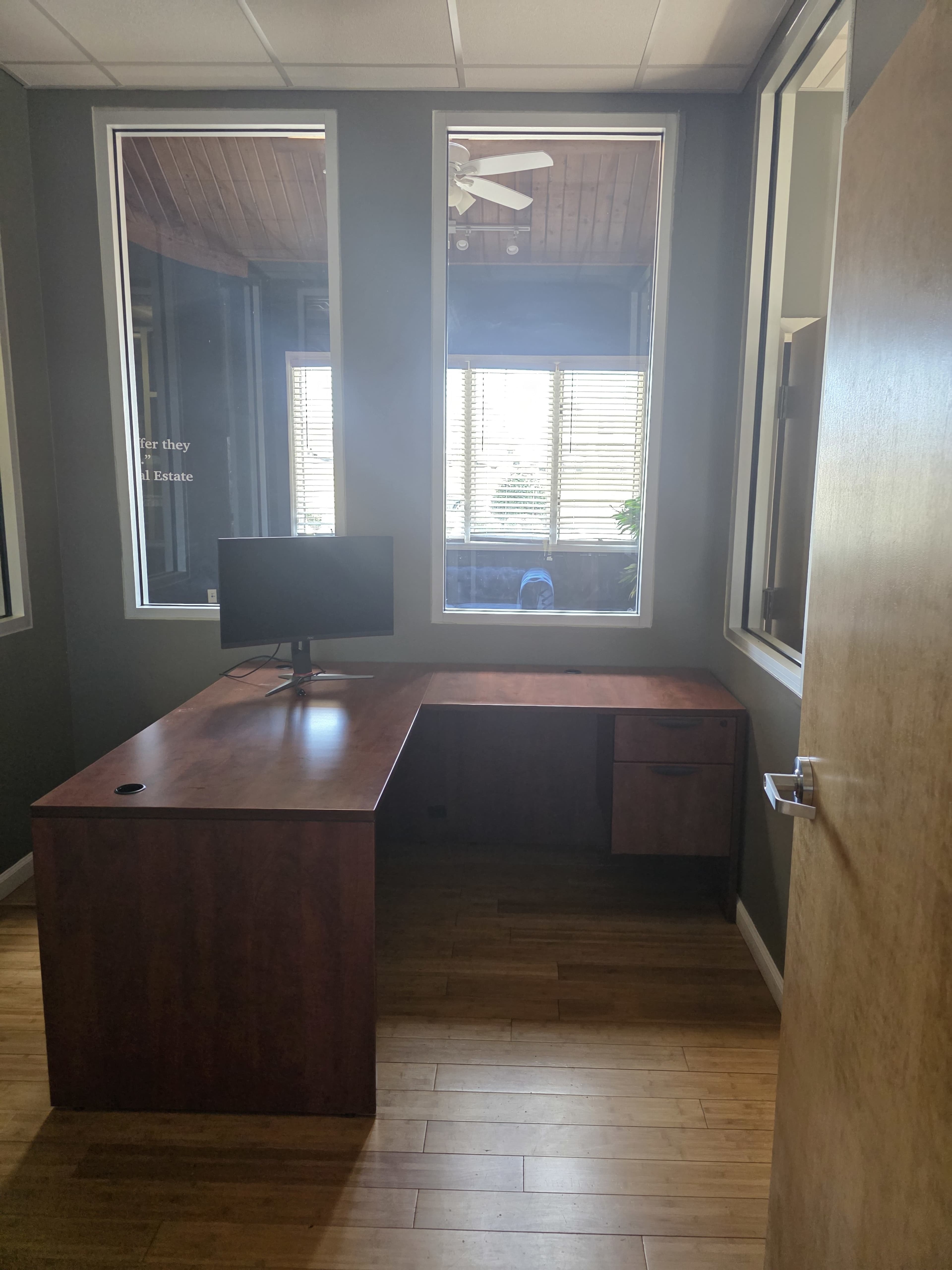 The image shows a modern office space featuring a wooden desk with a computer monitor, large windows allowing natural light, and bamboo flooring.
