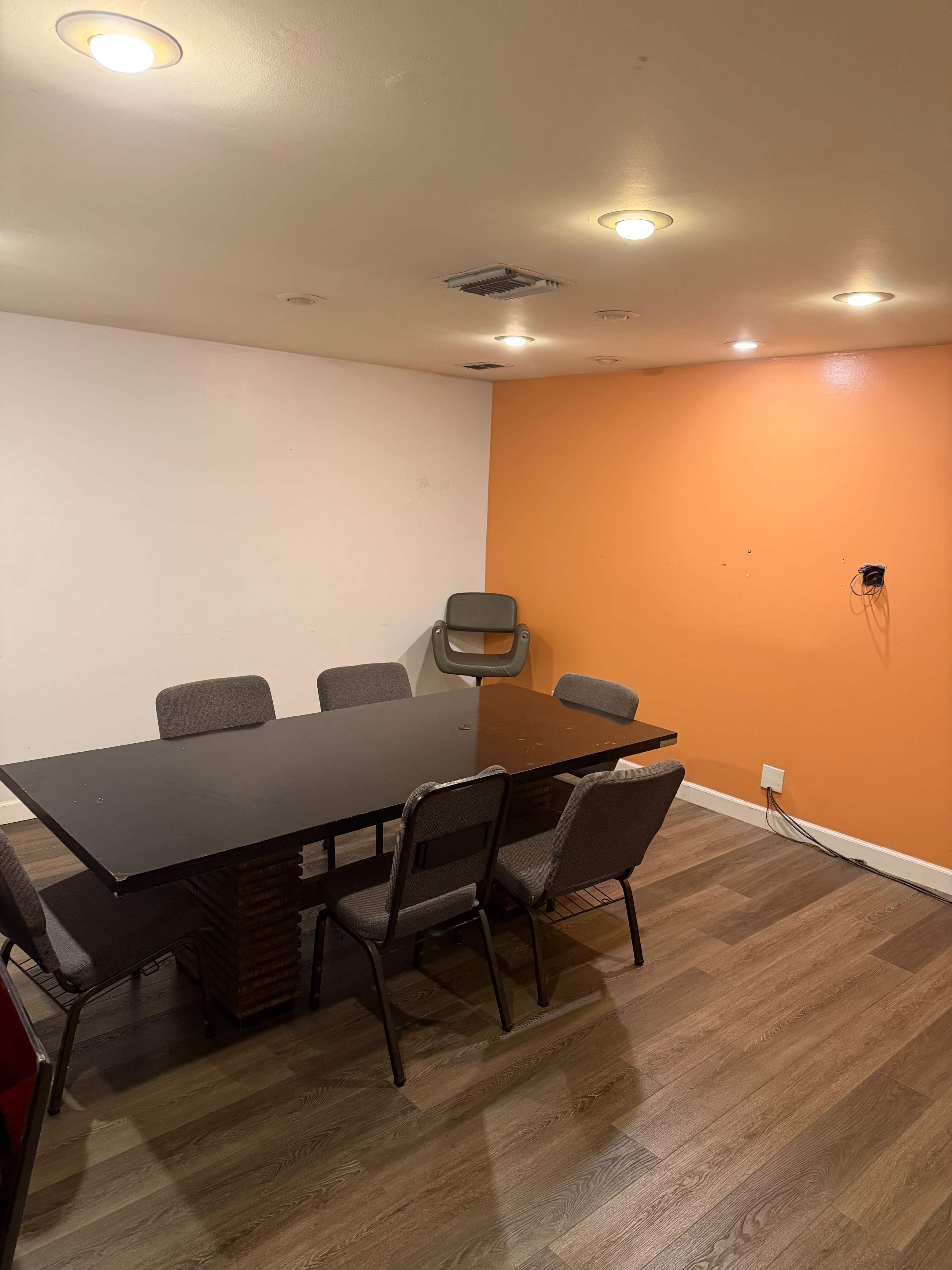 A room features a table surrounded by six chairs, with one chair positioned against an orange wall and the opposite wall painted white.