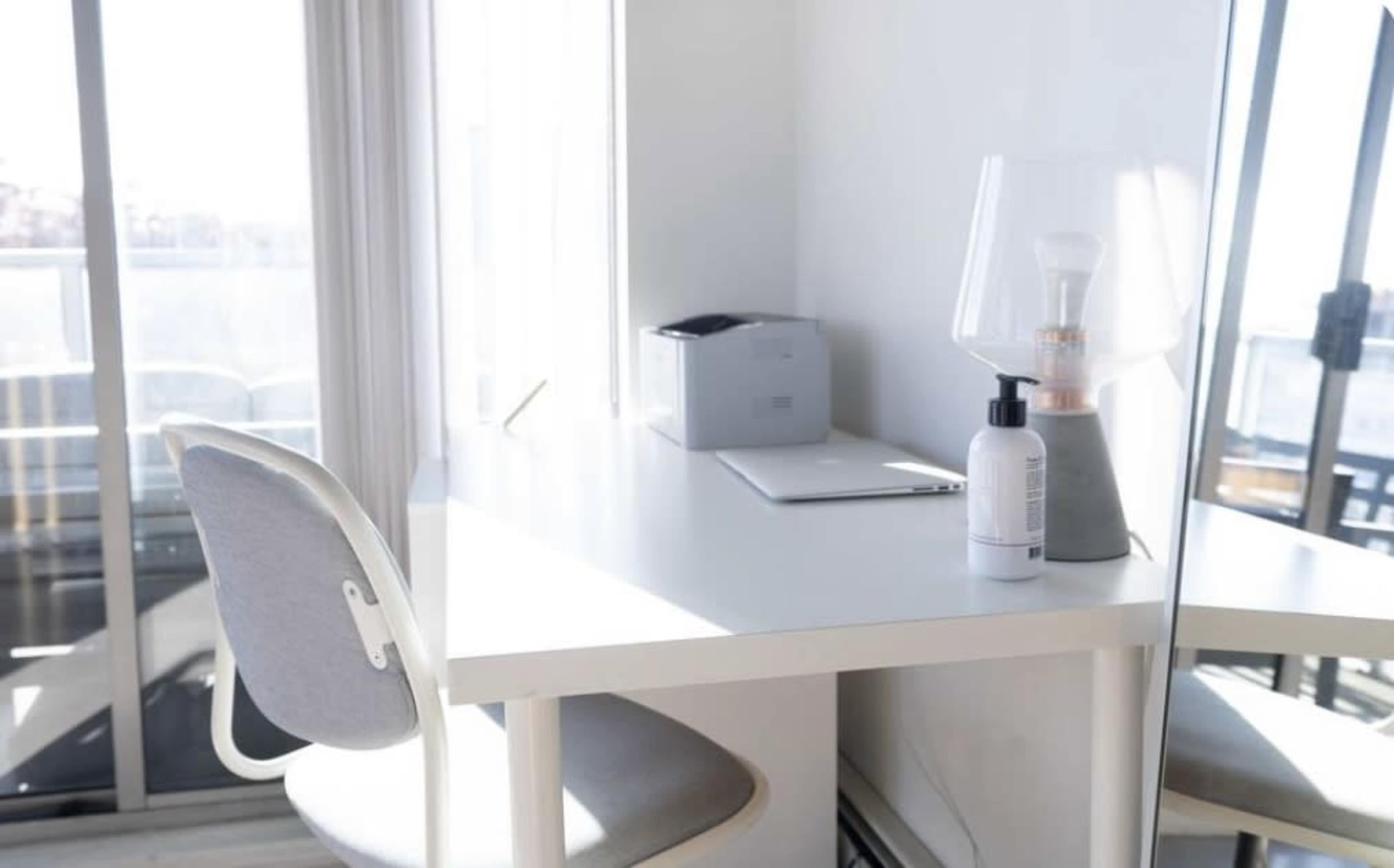 A minimalist workspace features a white desk, a laptop, a lamp, and a bottle on the surface, with large windows providing natural light.