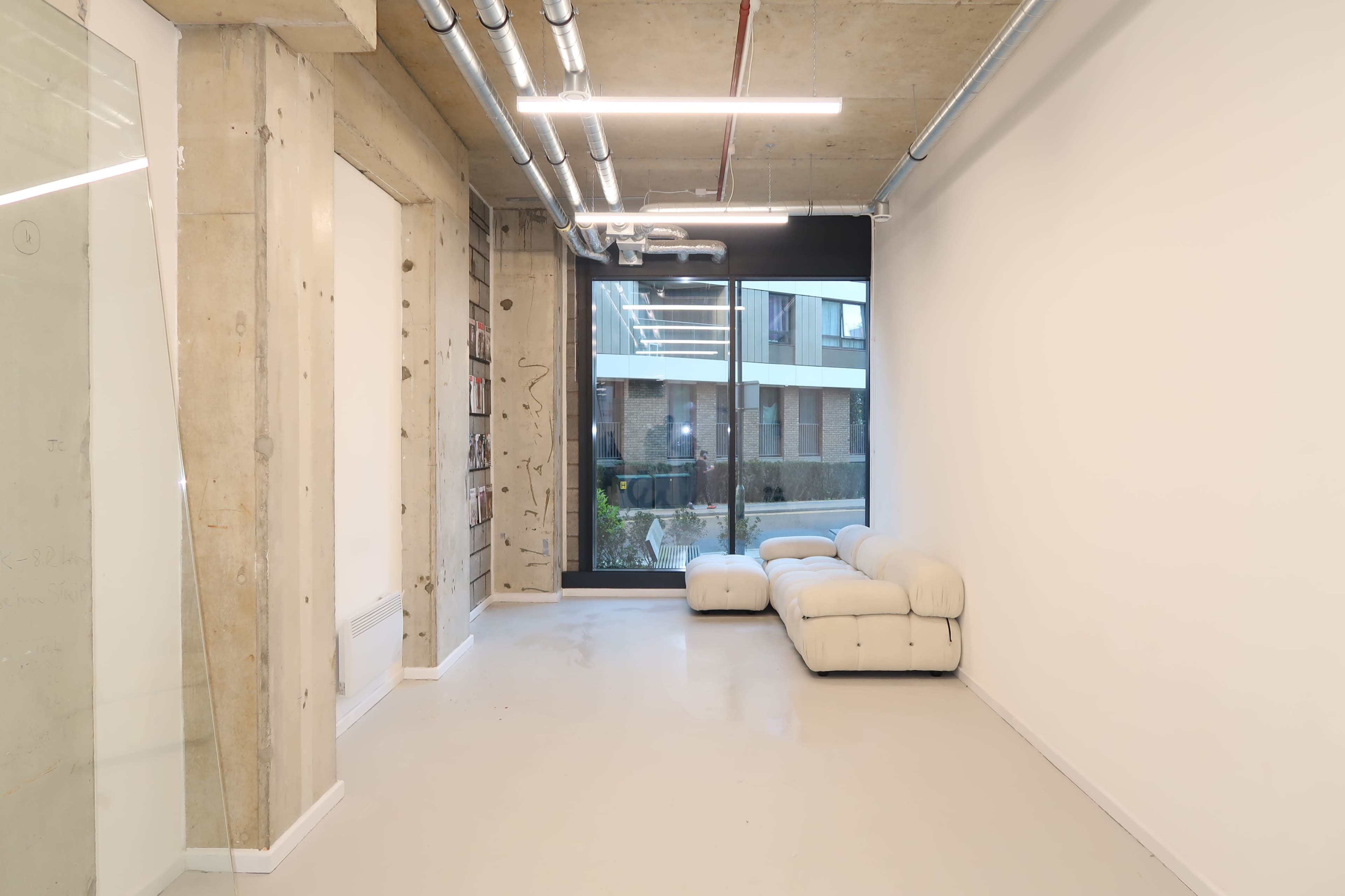 The image shows a minimalistic room featuring a white sofa, large window, and exposed concrete walls and ceiling.