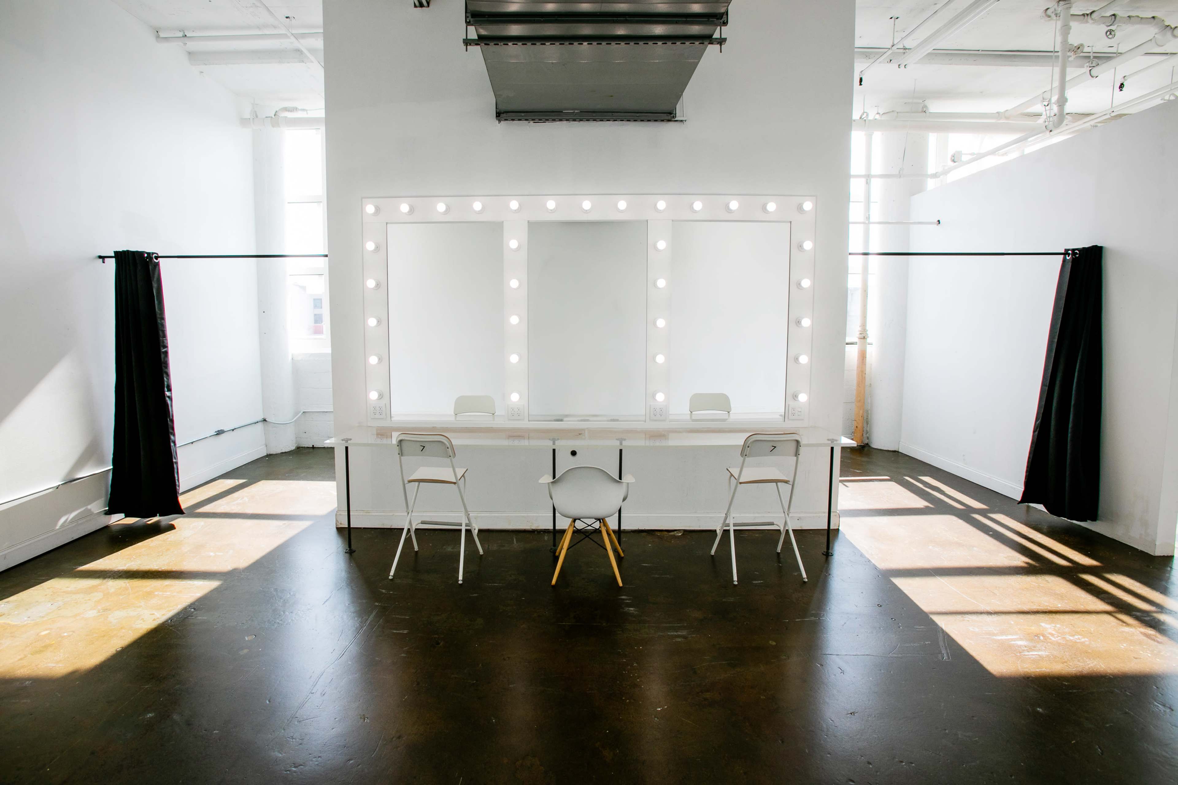 A brightly lit makeup room features a large mirror surrounded by lights, with a simple chair positioned in front and curtains hanging on either side.