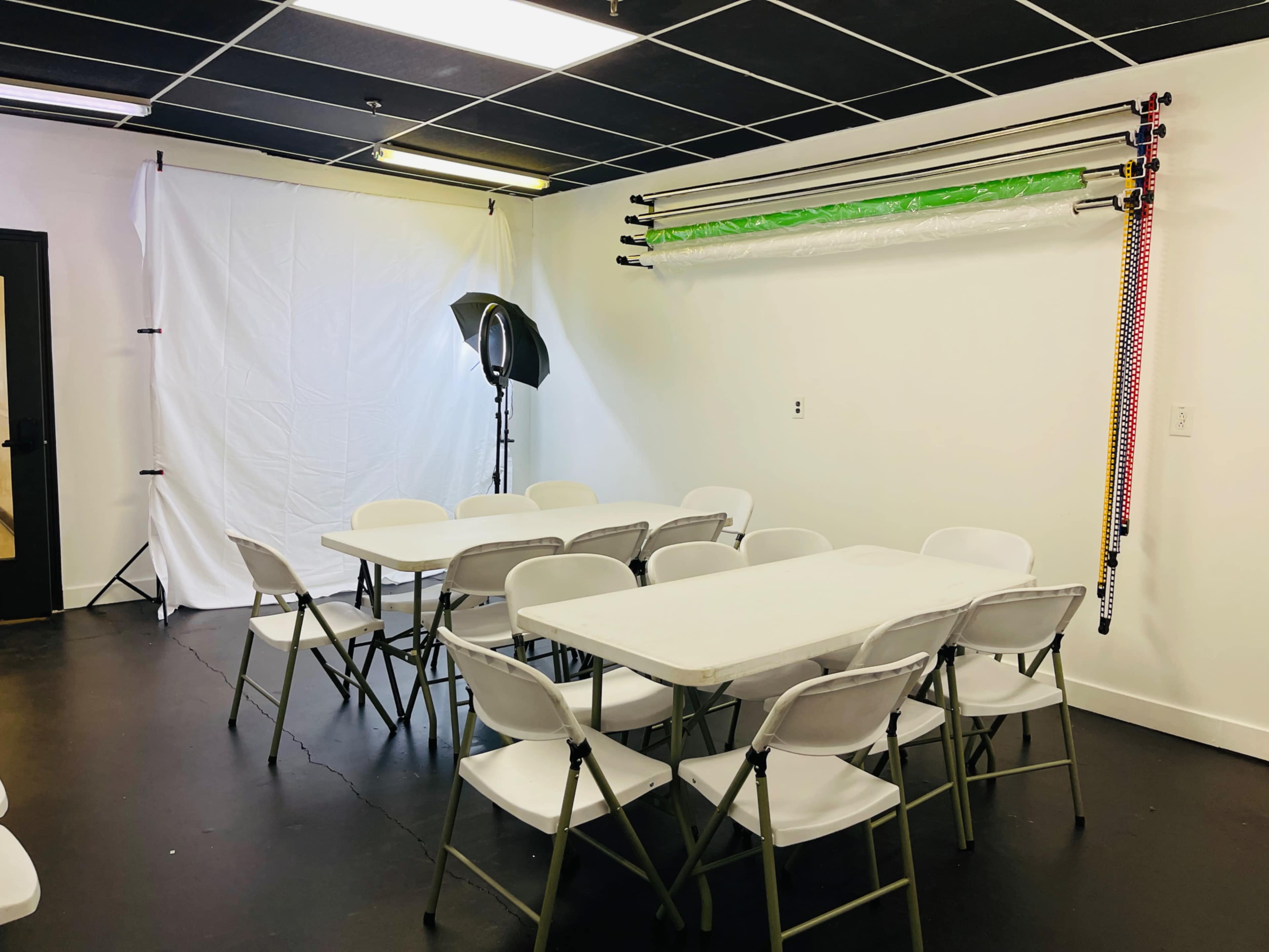 A meeting room with a white backdrop, a light source, and a row of folding chairs arranged around several tables.