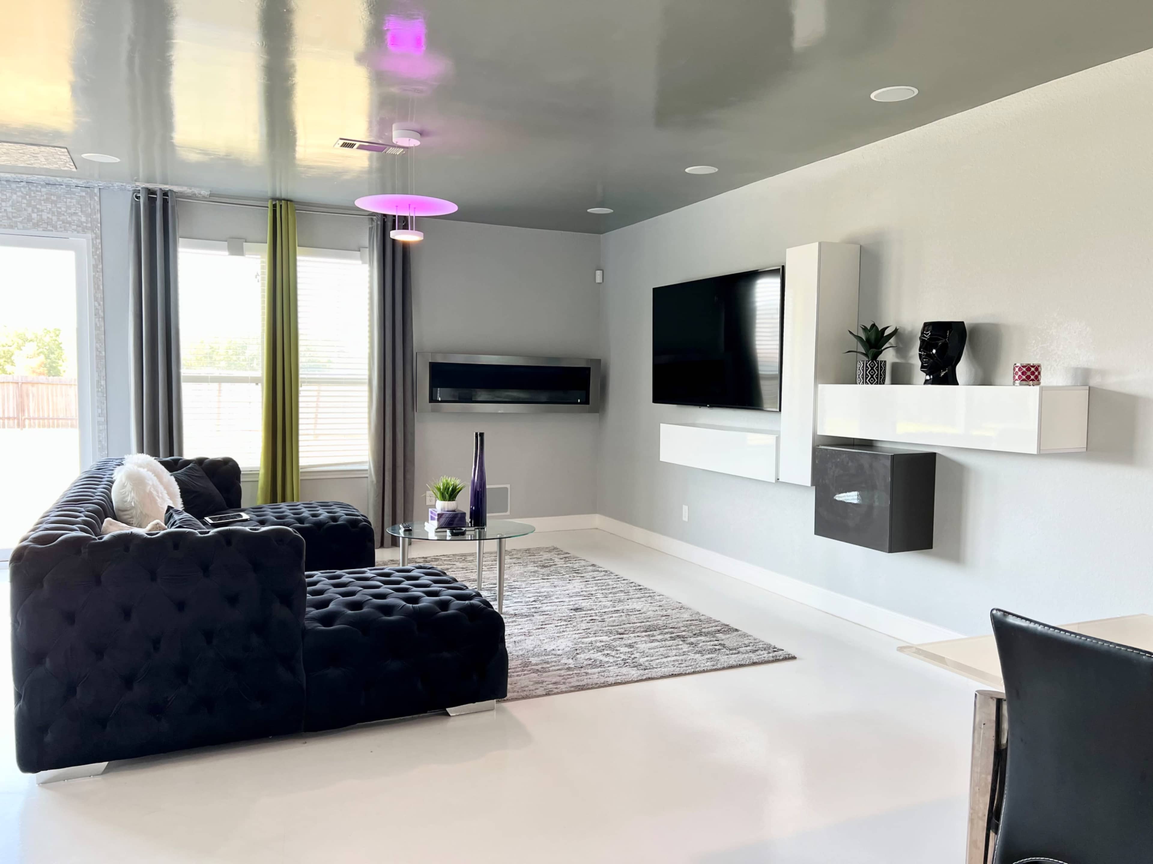 A modern living room with a black tufted sofa, a glass coffee table, and a wall-mounted television in a brightly lit space.