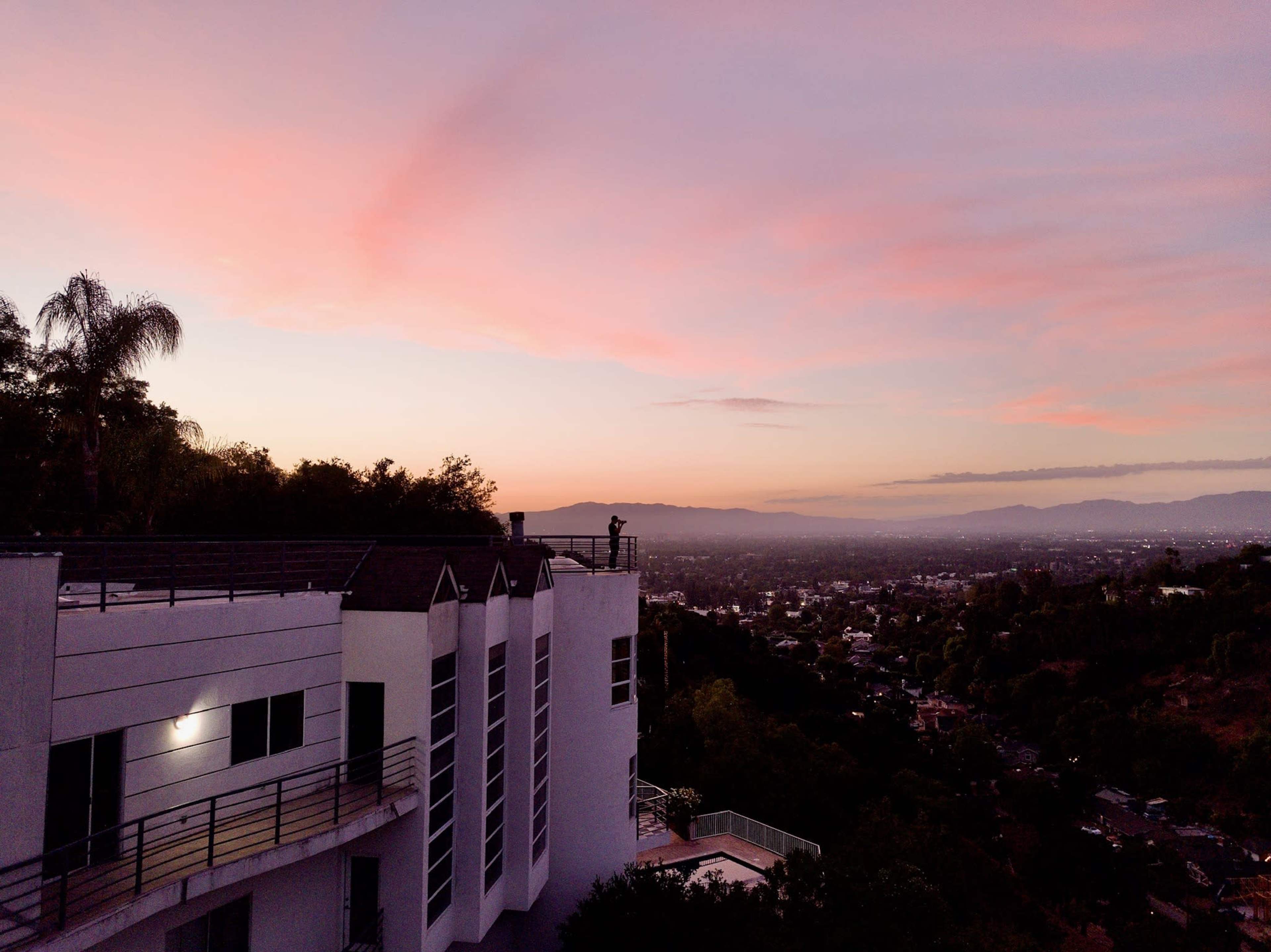 A hillside building overlooks a valley with a sunset-lit sky in the background.