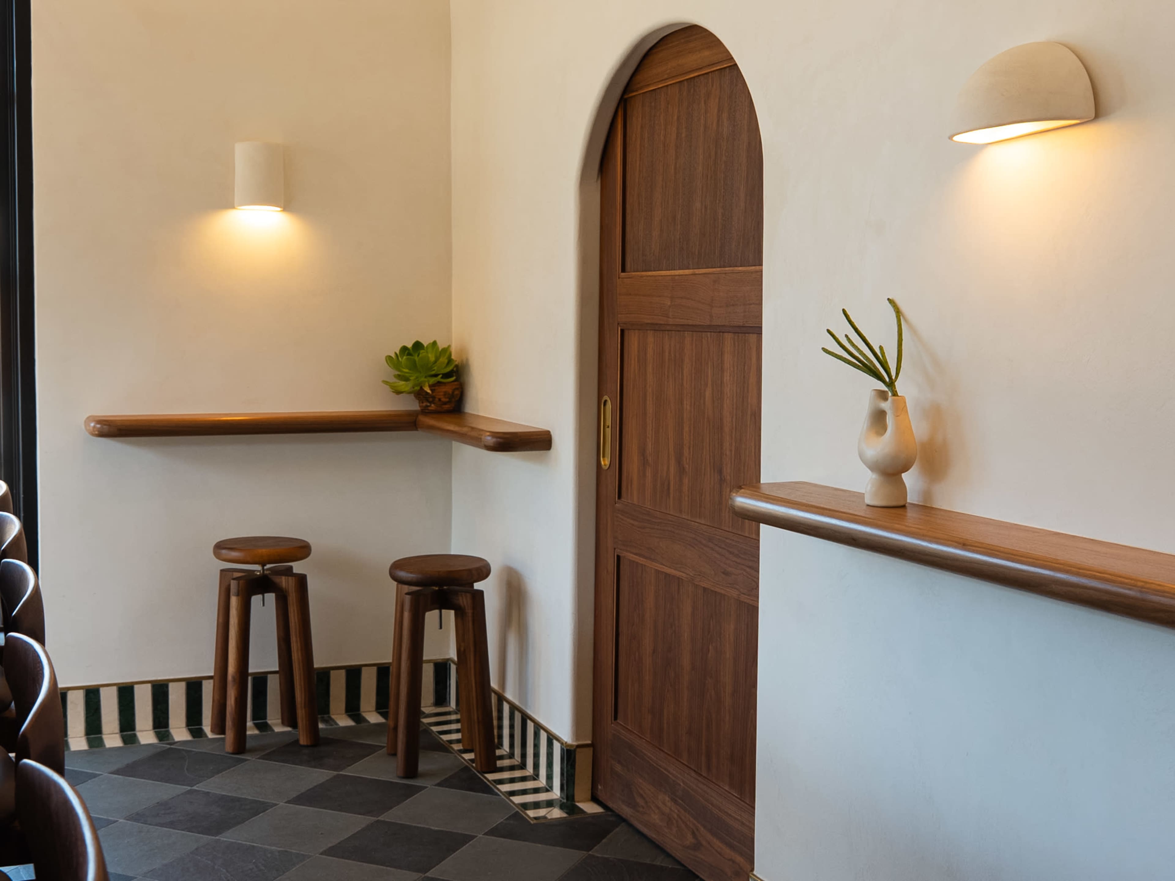 The image shows a minimalist interior with two wooden stools, a curved wooden wall shelf, and a simple door, complemented by neutral-colored walls and striped flooring.