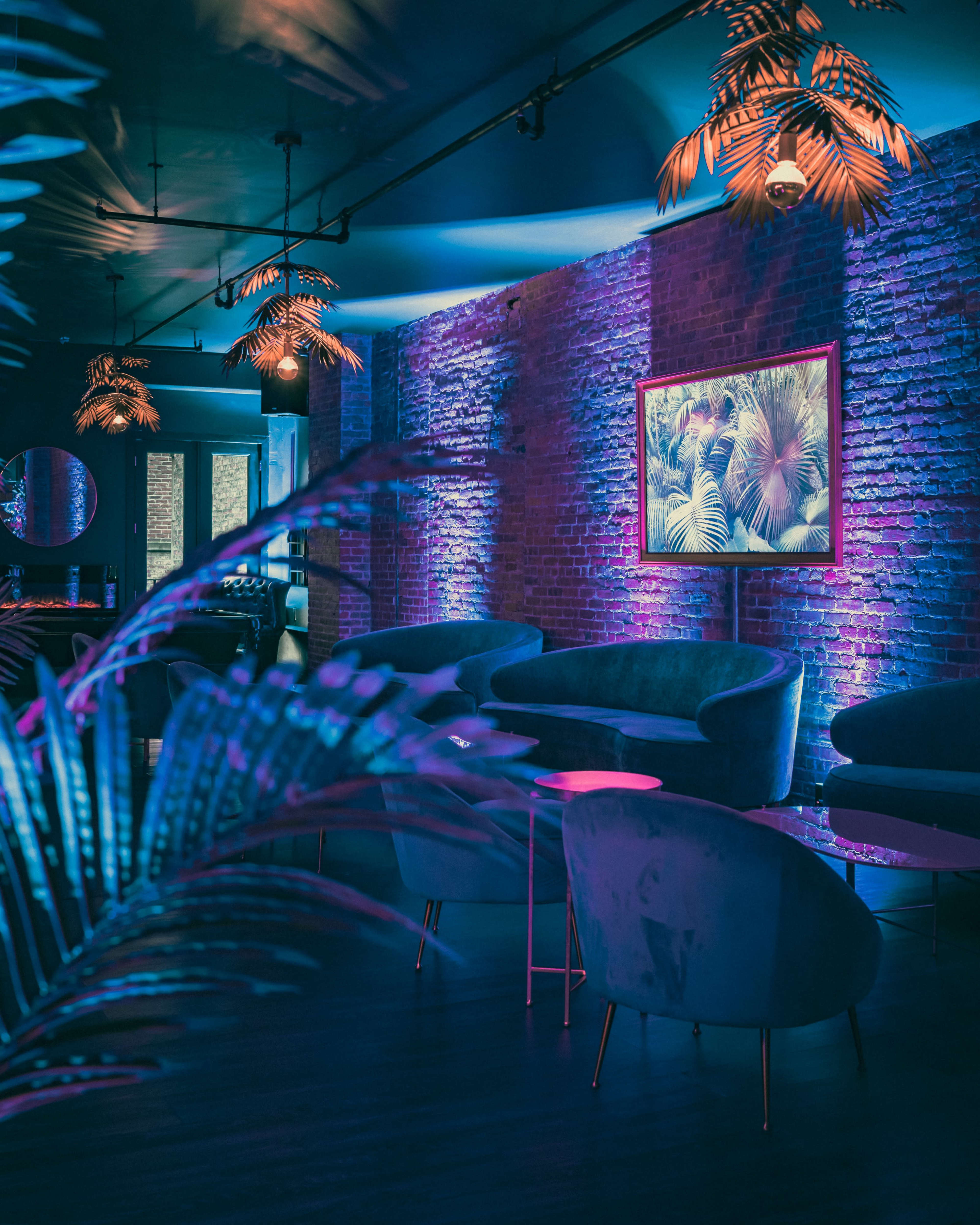 A modern lounge features plush seating, vibrant lighting, and a tropical-themed artwork framed on a brick wall.