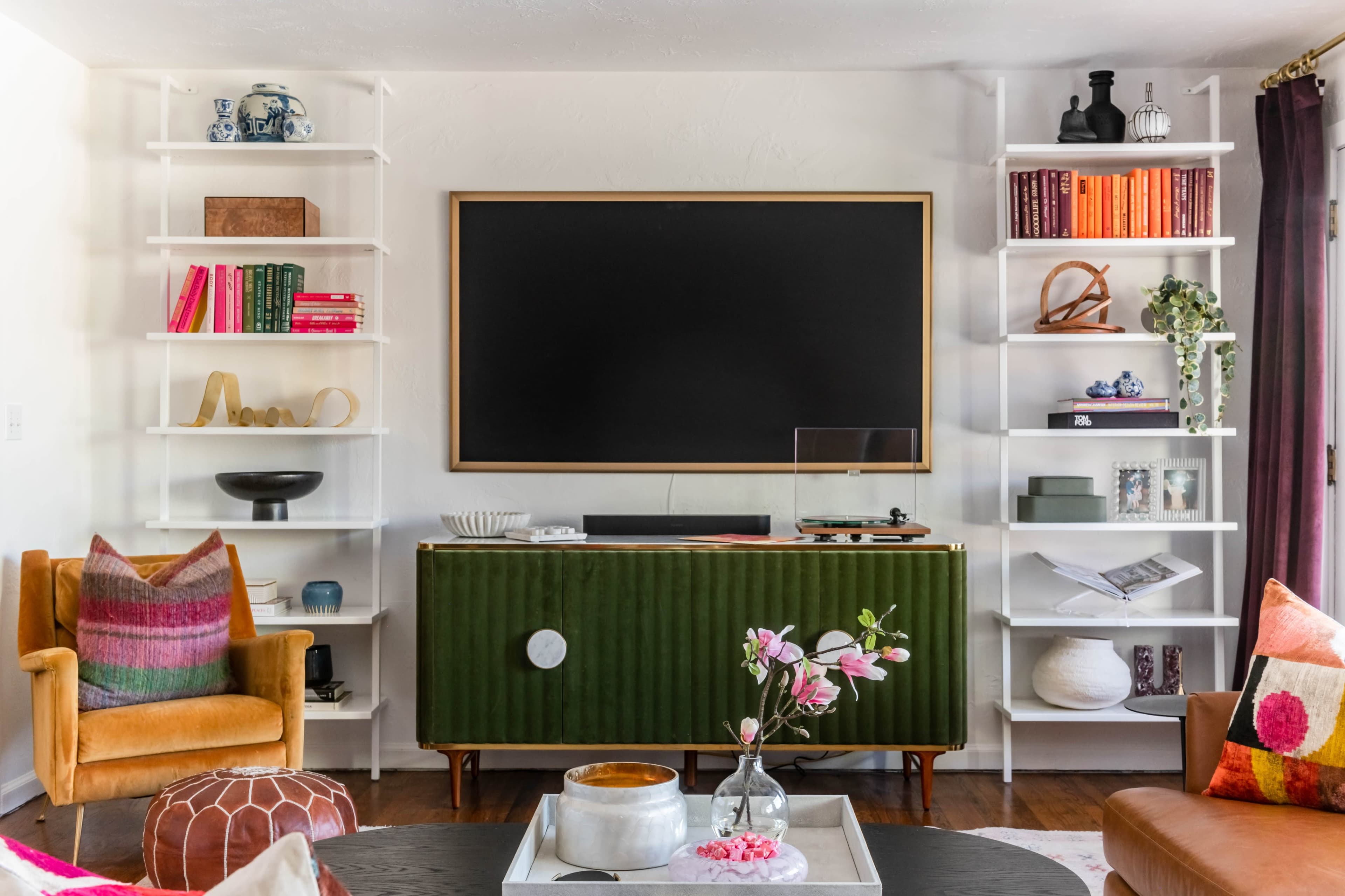 The image depicts a living room with a green media console, a large flat-screen TV mounted above it, and white shelving units filled with various decorative items and books.