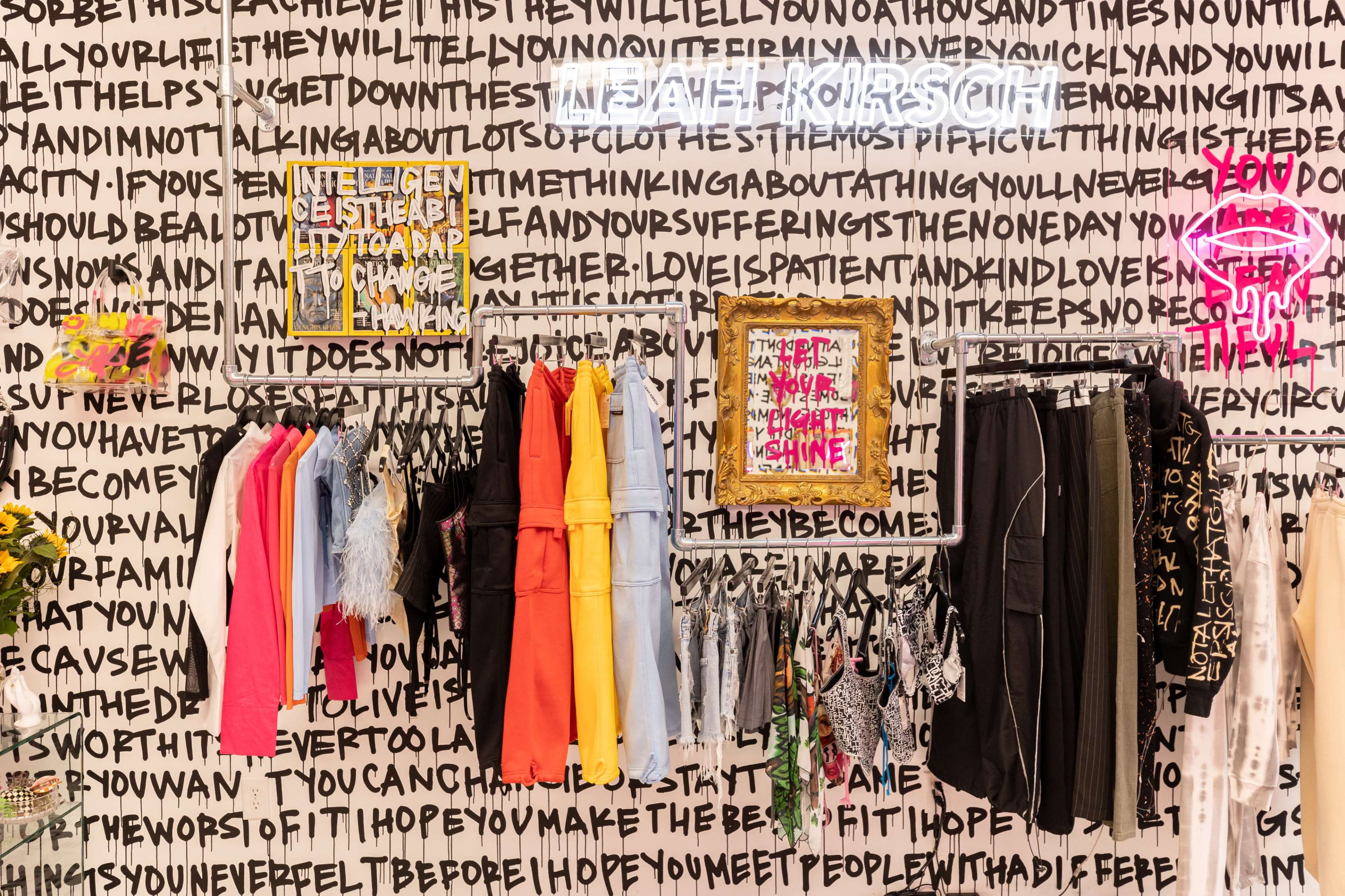 A clothing rack displaying various colorful garments against a wall covered in bold, black text and graphic art.