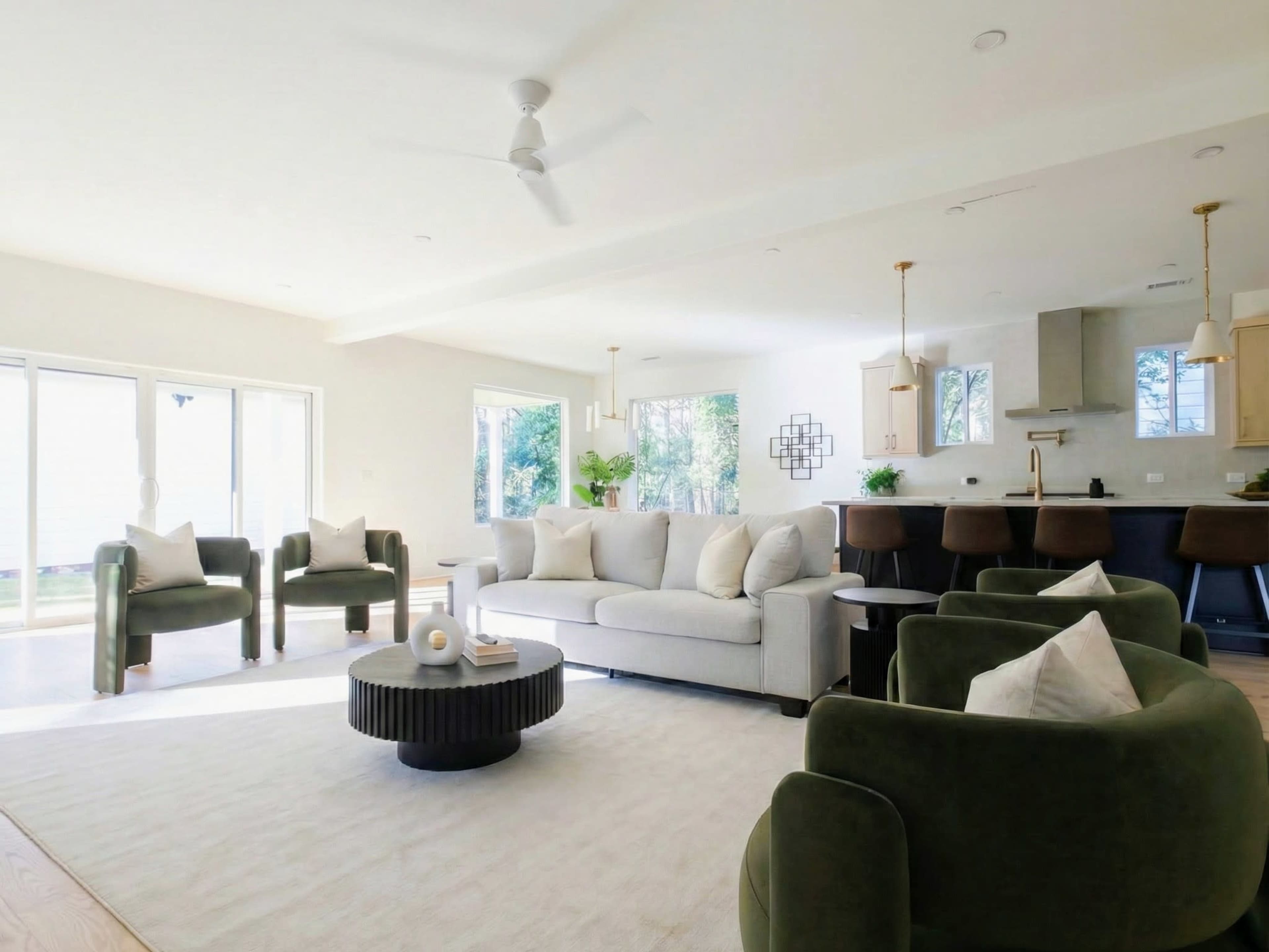The image shows a modern living room with a light-colored sofa, two green armchairs, a round coffee table, and an open kitchen area in the background.