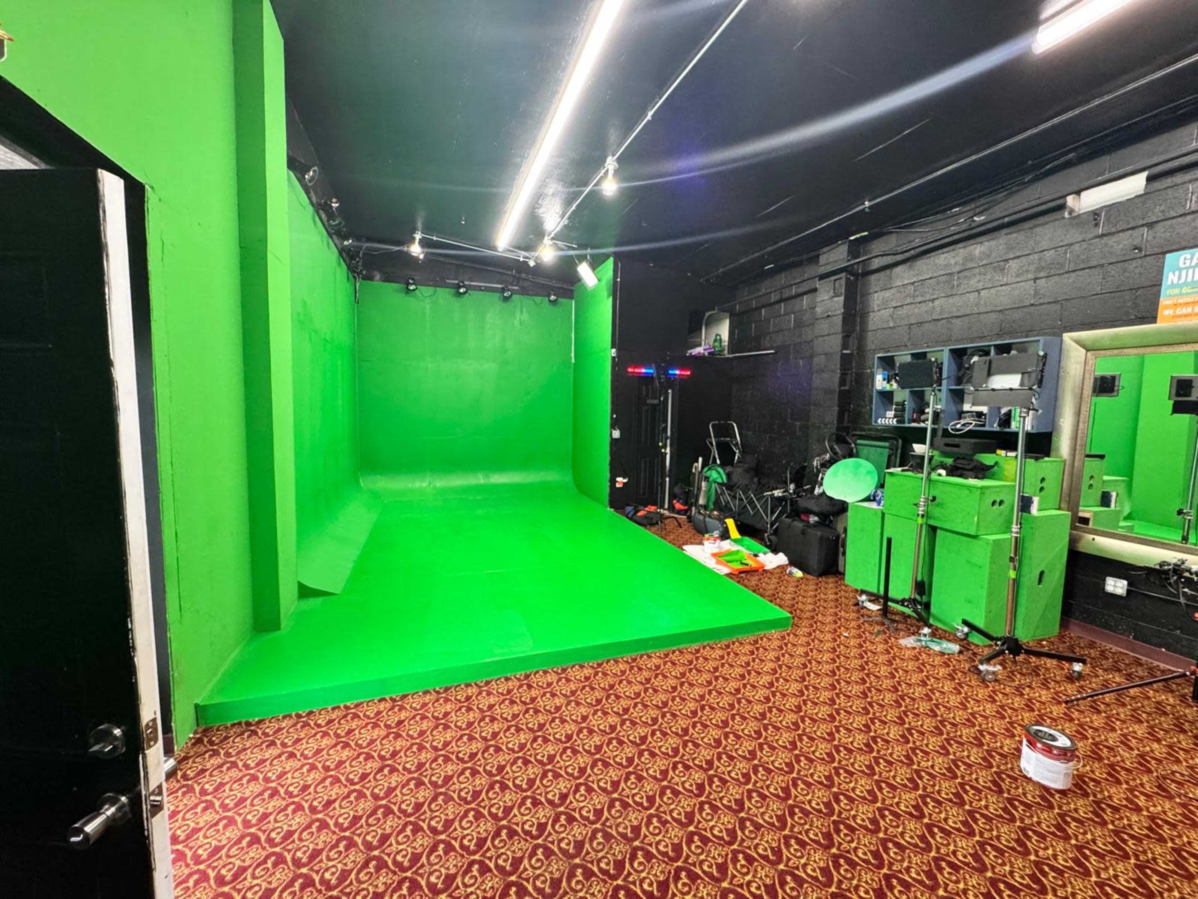 The image shows a green screen setup in a room with black walls and a patterned carpet, along with various equipment and clutter.