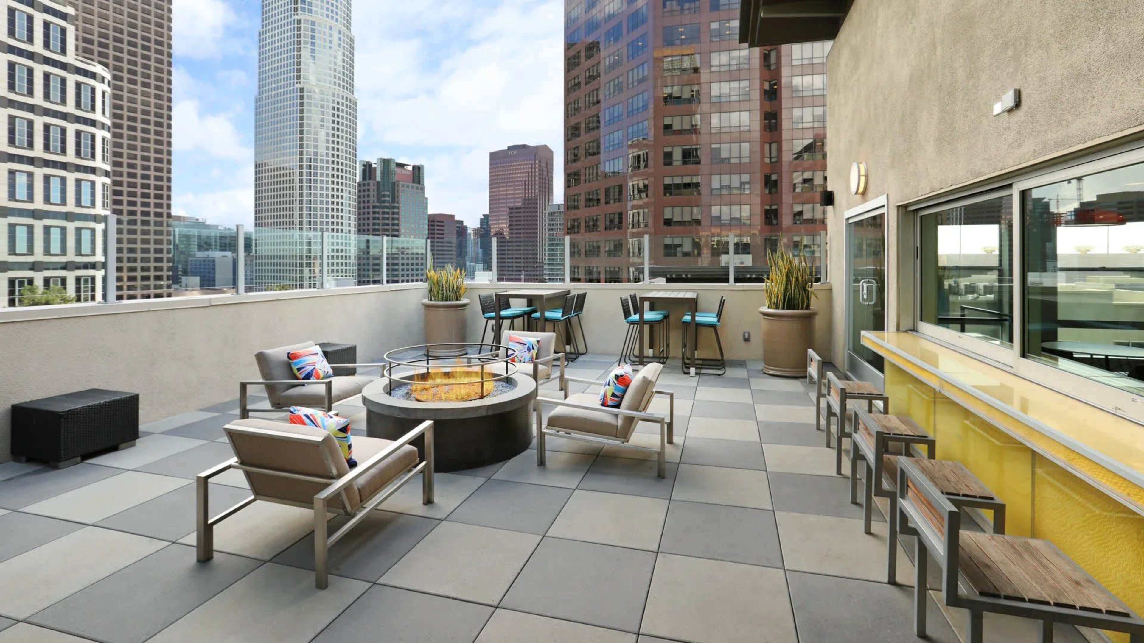 A rooftop terrace featuring modern seating around a circular fire pit, with tall buildings and a clear sky in the background.