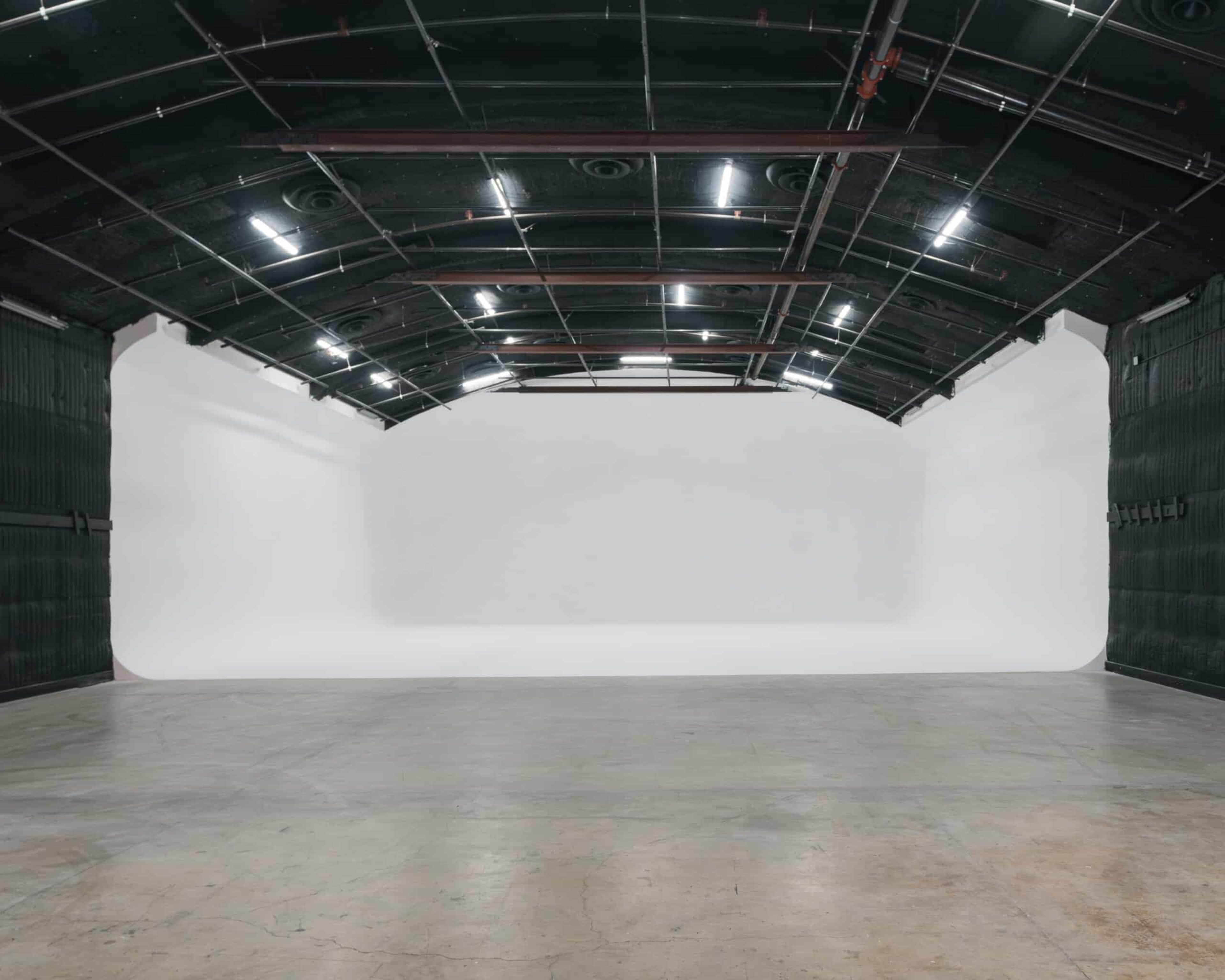 The image shows a spacious, empty studio with a large white backdrop and high, dark green ceilings.