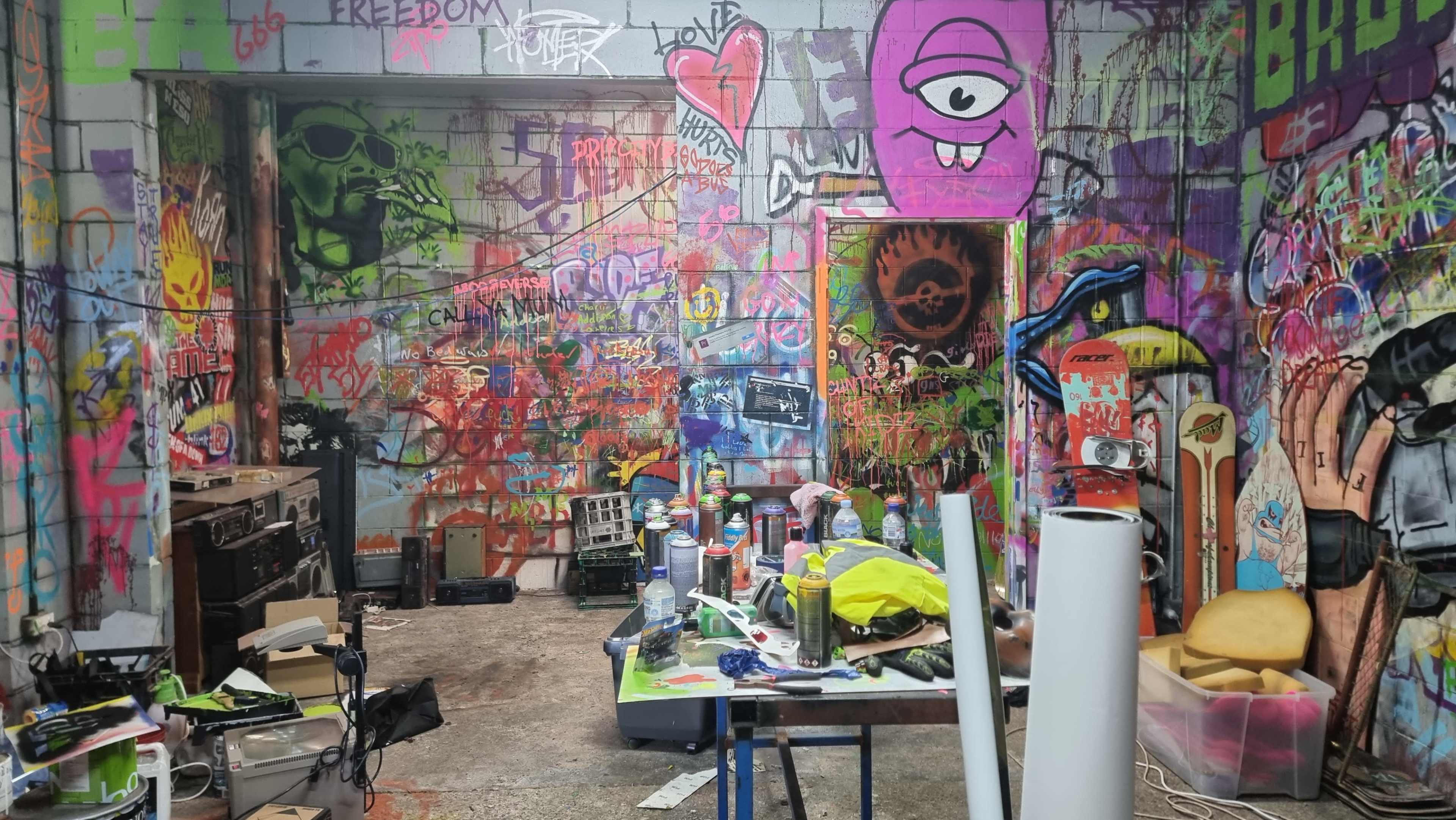 A cluttered indoor space filled with colorful graffiti on the walls and various art supplies scattered around a table.