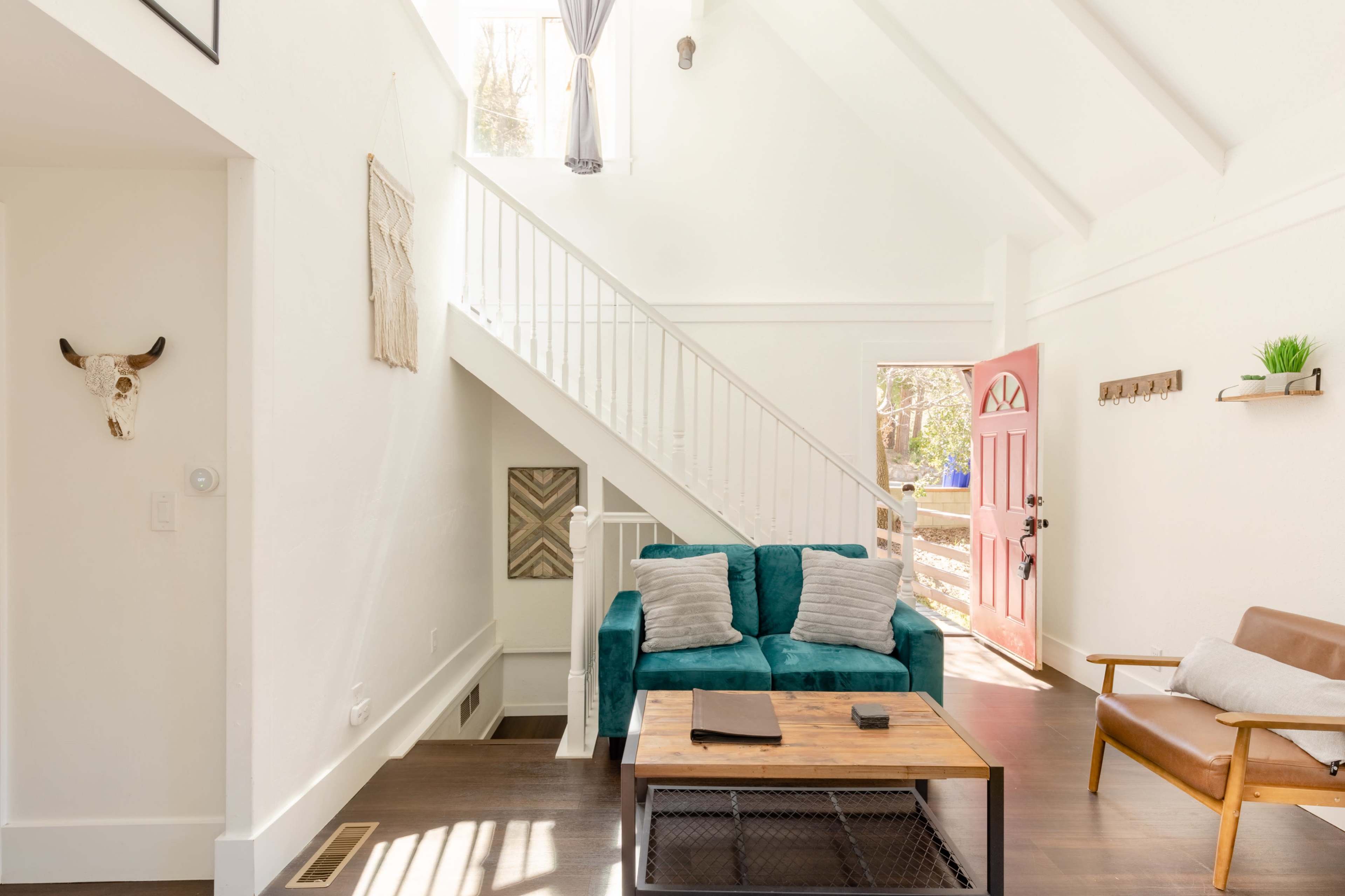 The image shows a bright interior with a teal sofa, wooden coffee table, and a staircase leading to an upper level, with a pink door open to a view outside.